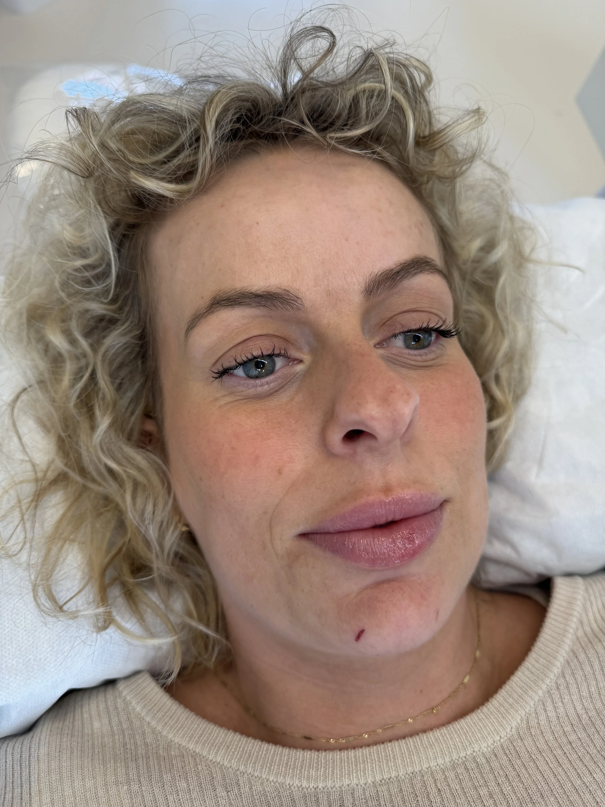 Nærbillede af en kvindes ansigt med lyse, krøllede hår, blå øjne, let makeup, ferskenfarvet læbestift, en enkel guldhalskæde og en beige sweater.