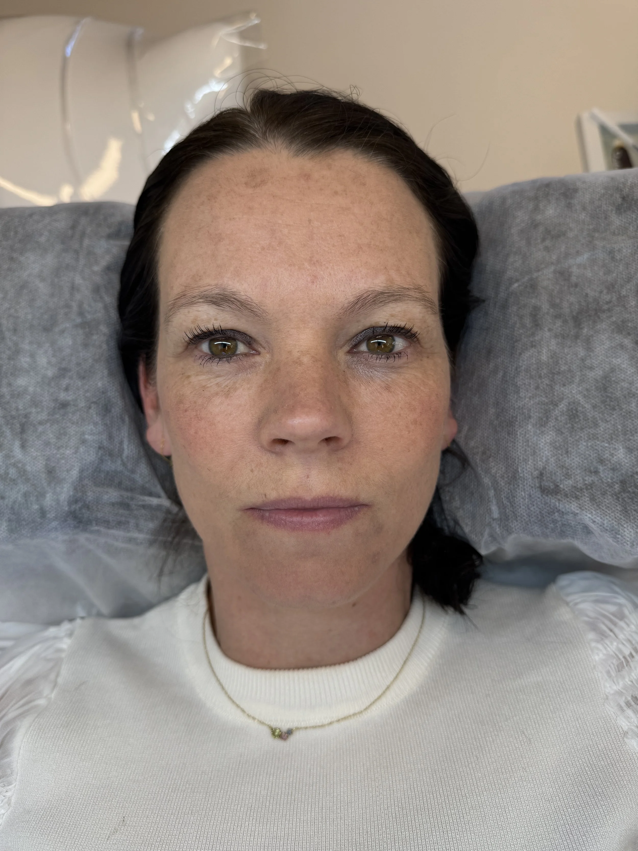 Nærbillede af en kvinde, der ligger ned med hovedet på en pude, har brunt hår, grønne øjne og bærer en hvid trøje og en halskæde.