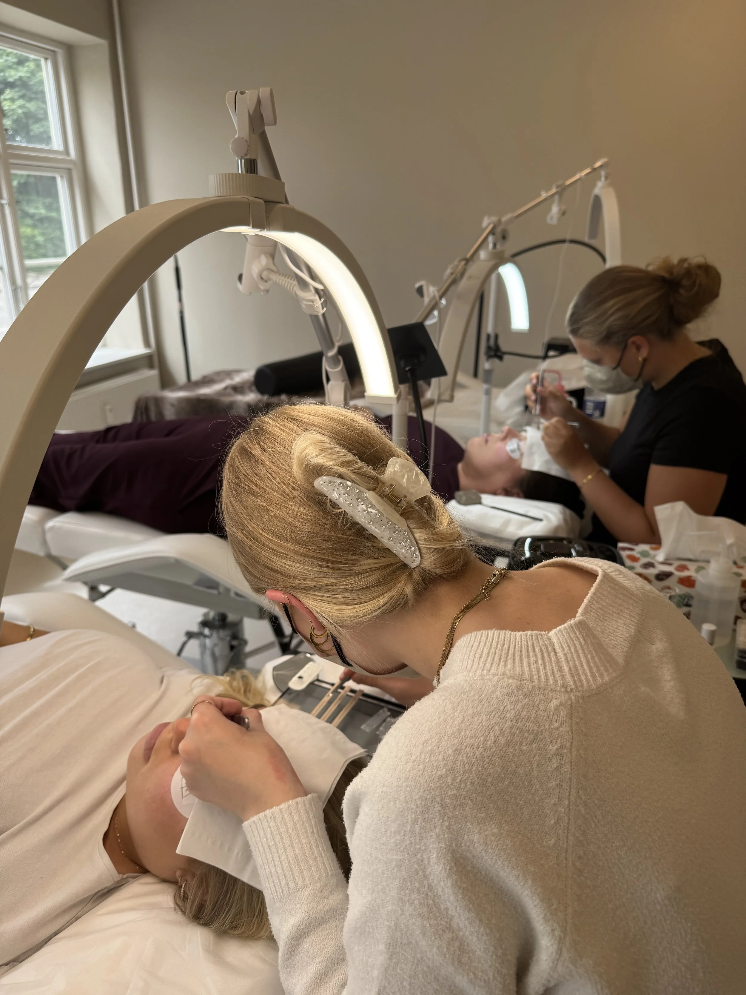 En person med blondt hår og hårbøjle arbejder på en skønhedsbehandling på en anden person, der ligger på en klinik med medicinsk udstyr og en person med ansigtsmaske.