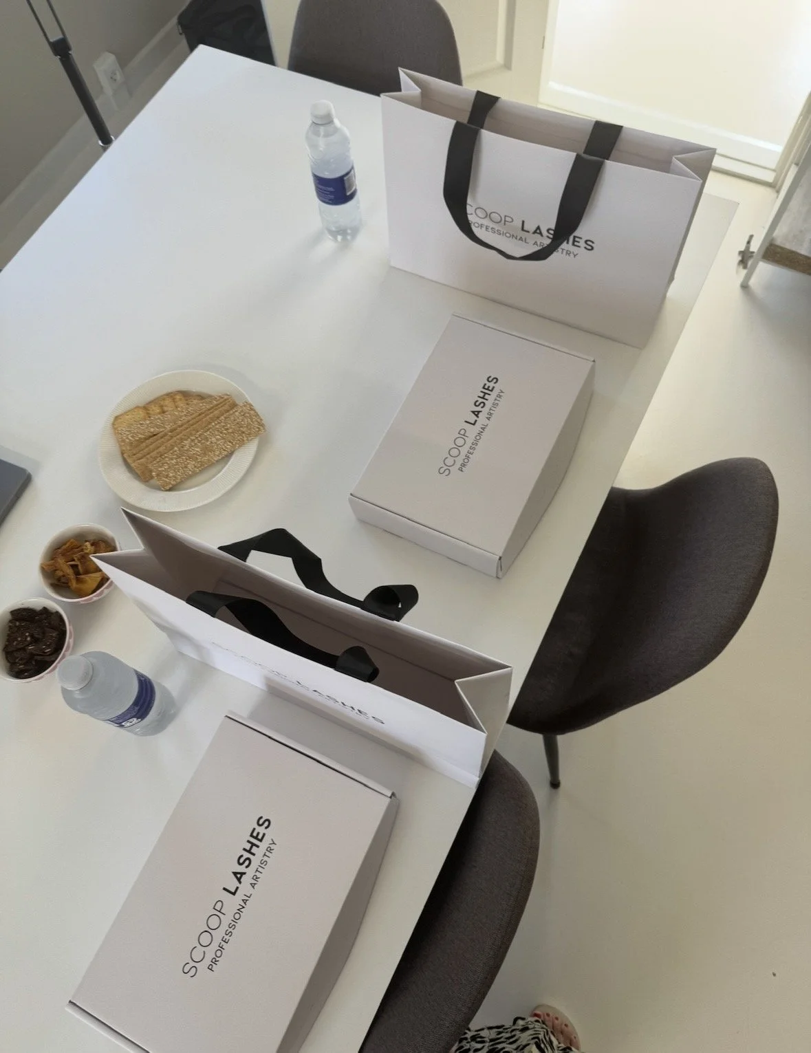 En hvid bord med adskillige koffertæsken med logoet 'SCOOP LASHES', en vandflaske, en tallerken med sprød snacks, og tre små skåle med tørret frugt og chokolade. To stole fylder pladsen, og der er en stor papirpose på bordet og et vindue i baggrunden