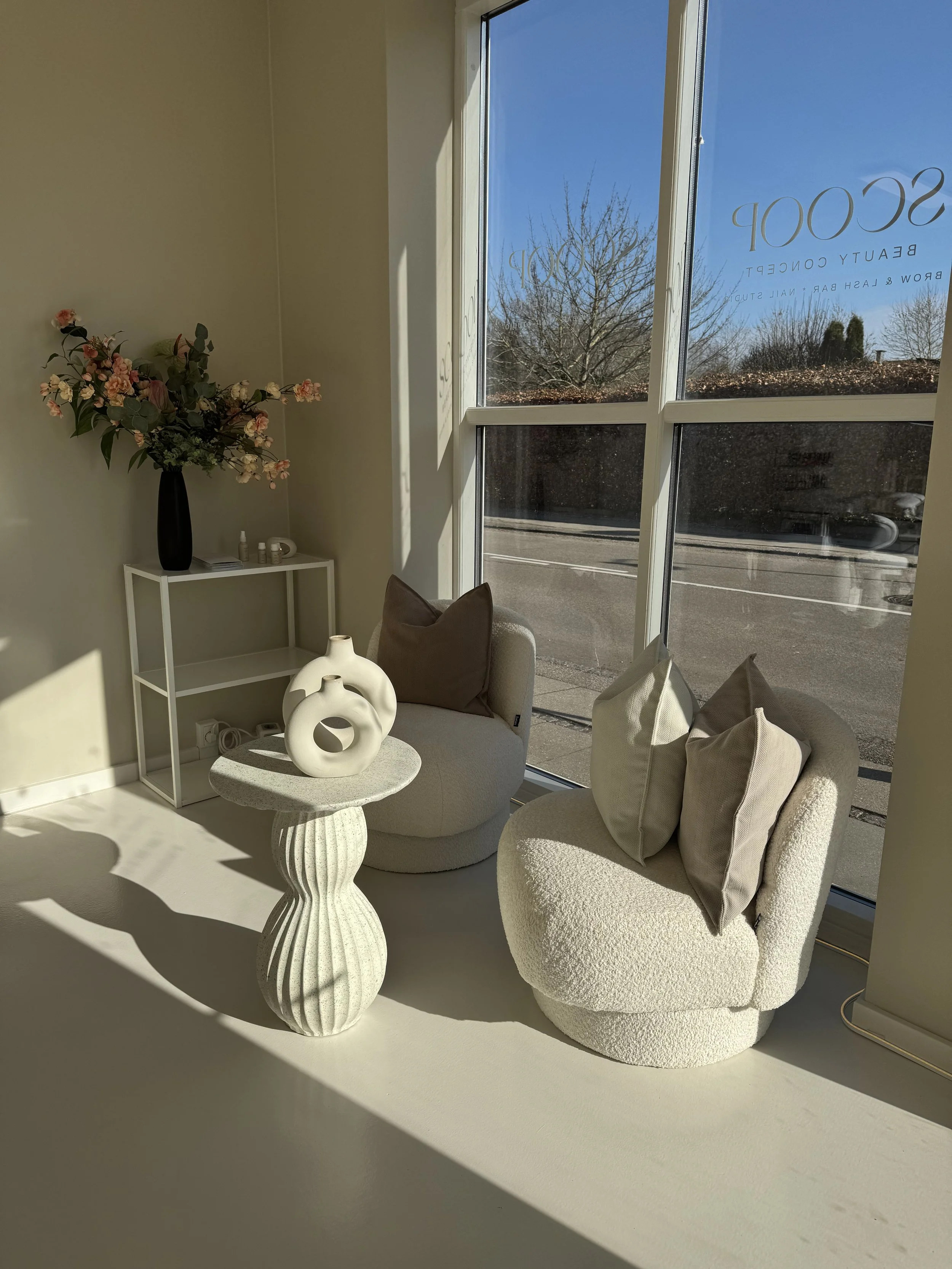 Indendørs: en hvid stol med beige pude, et hvidt rundt bord med to dekorative hvide vaser, en hvid reol med en sort vase med blomster og nogle små dekorative ting, store vinduer med udsyn til en blå himmel og træer uden blade.