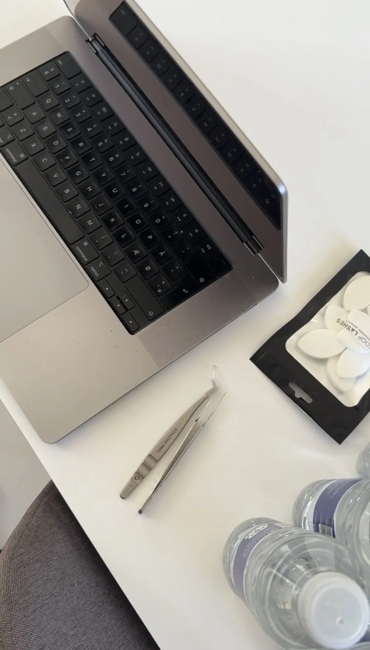 Billedet viser en laptop, medicin, store vandflasker, en pen, pincet og nogle tabletter på et skrivebord.