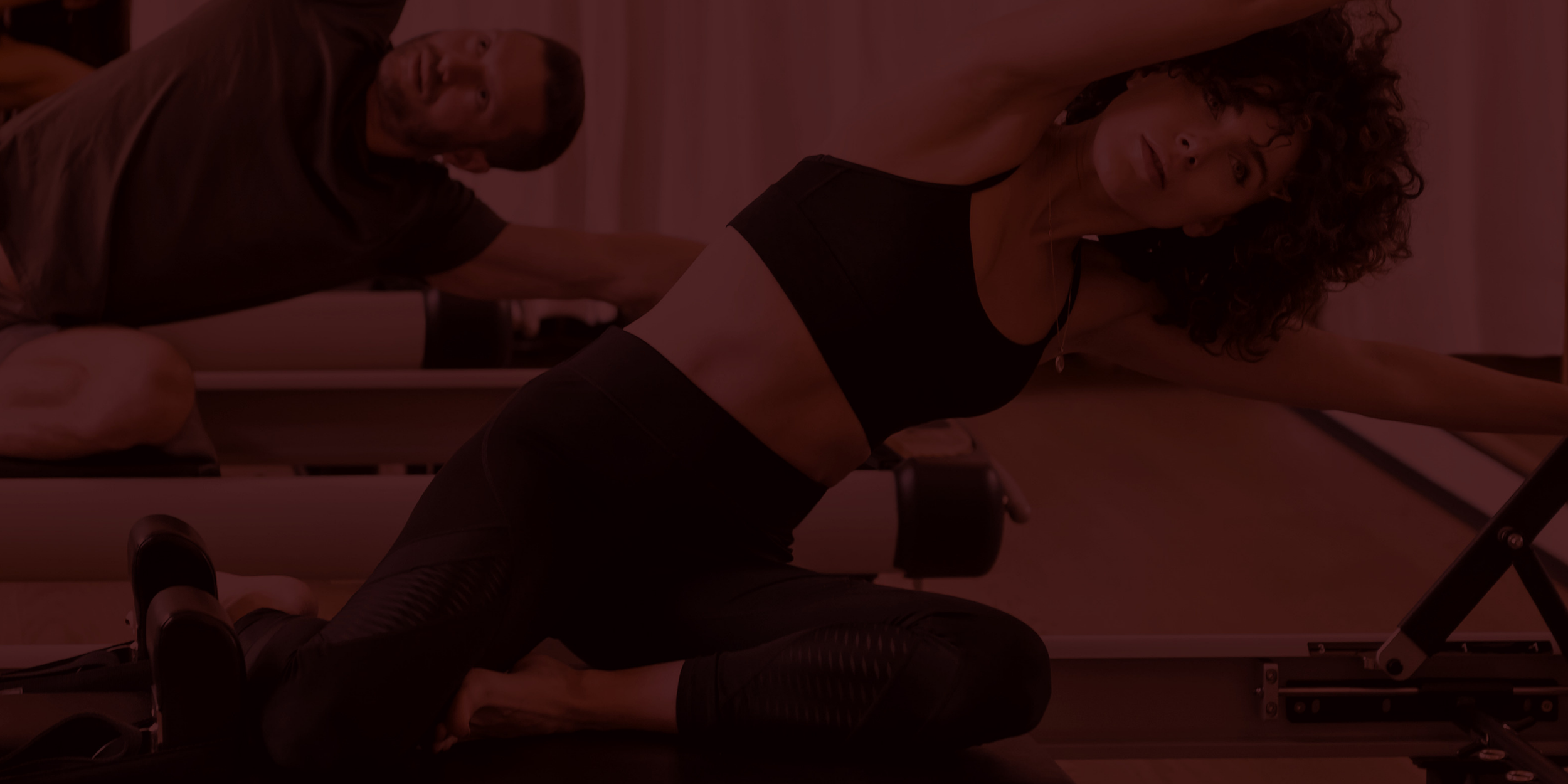 A woman in athletic wear doing a yoga pose or stretch on a reformer machine, with a man in the background observing or assisting.