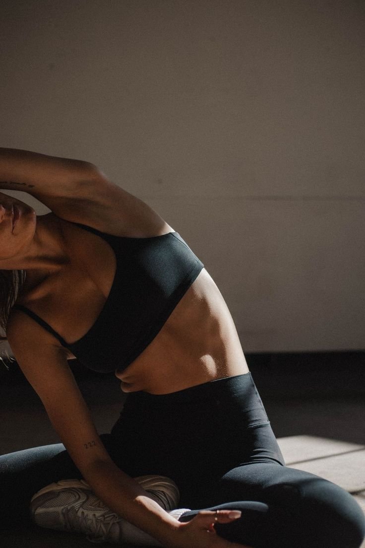 A woman in black sportswear performs a stretching exercise indoors on a dark floor with sunlight coming through a window.