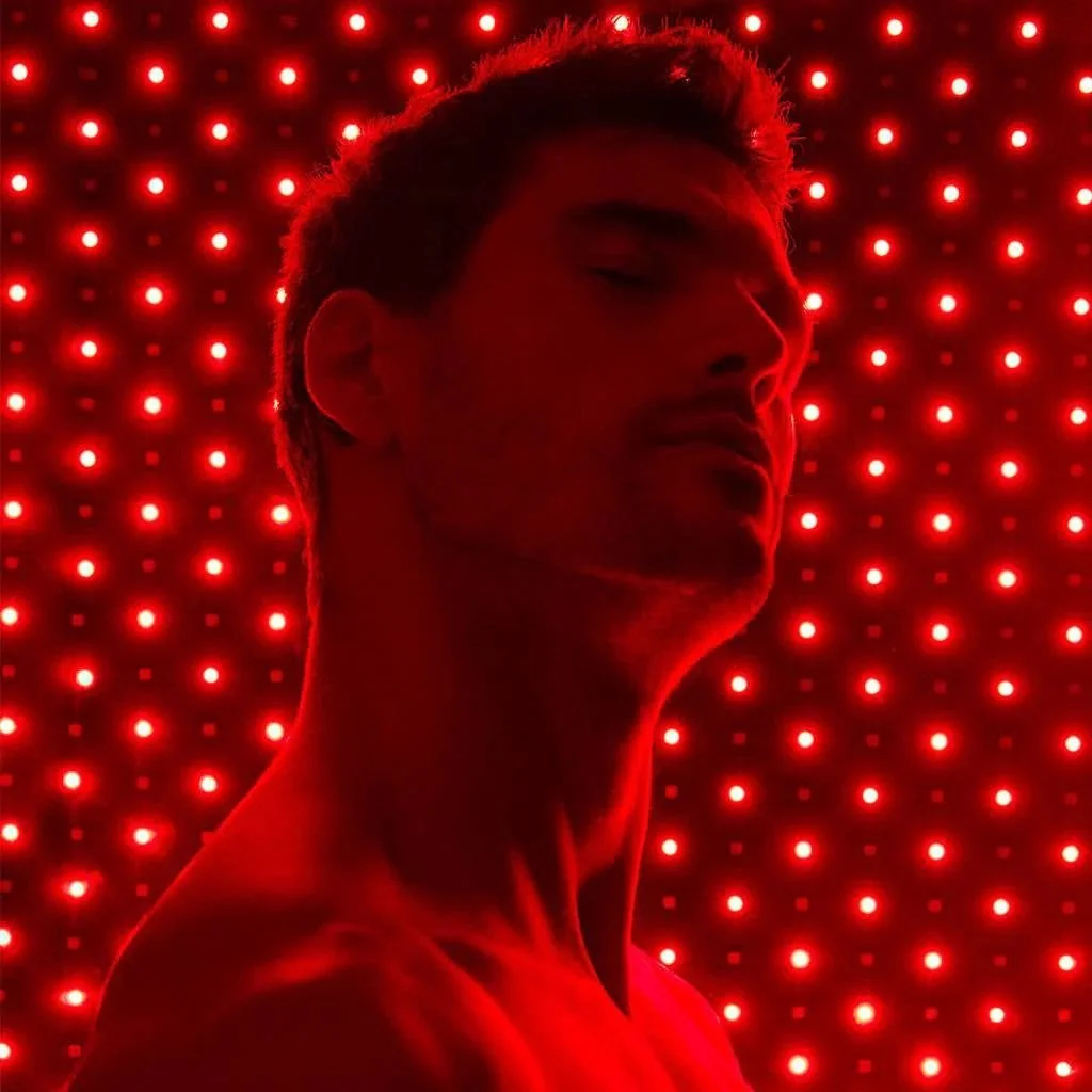 A shirtless man with short hair stands in front of a wall of red LED lights, with his eyes closed and head tilted slightly upward.