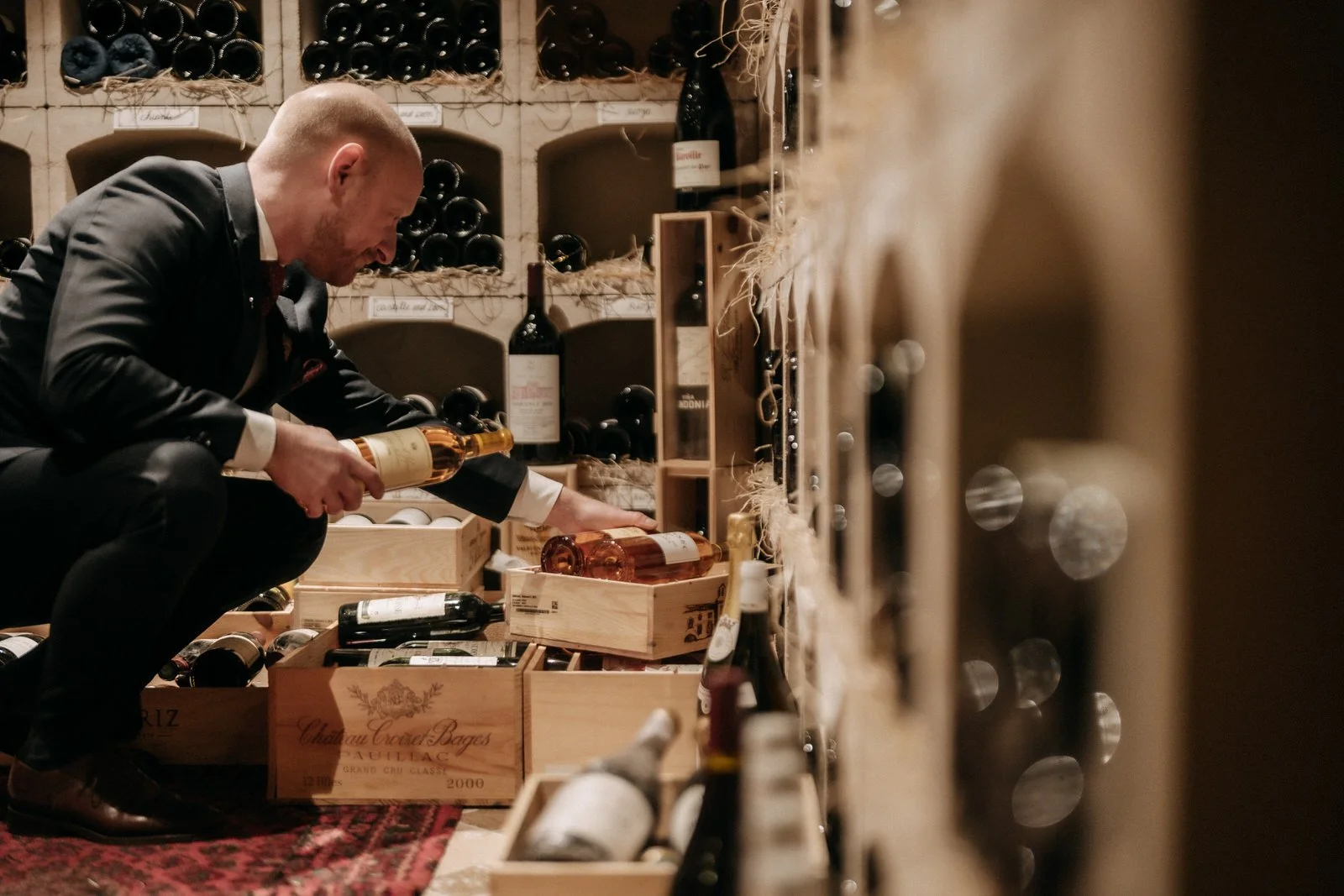 A sommelier in a suit is exploring the wine cellar of Williams Farmhouse, examining bottles of wine stored in wooden boxes and shelves.