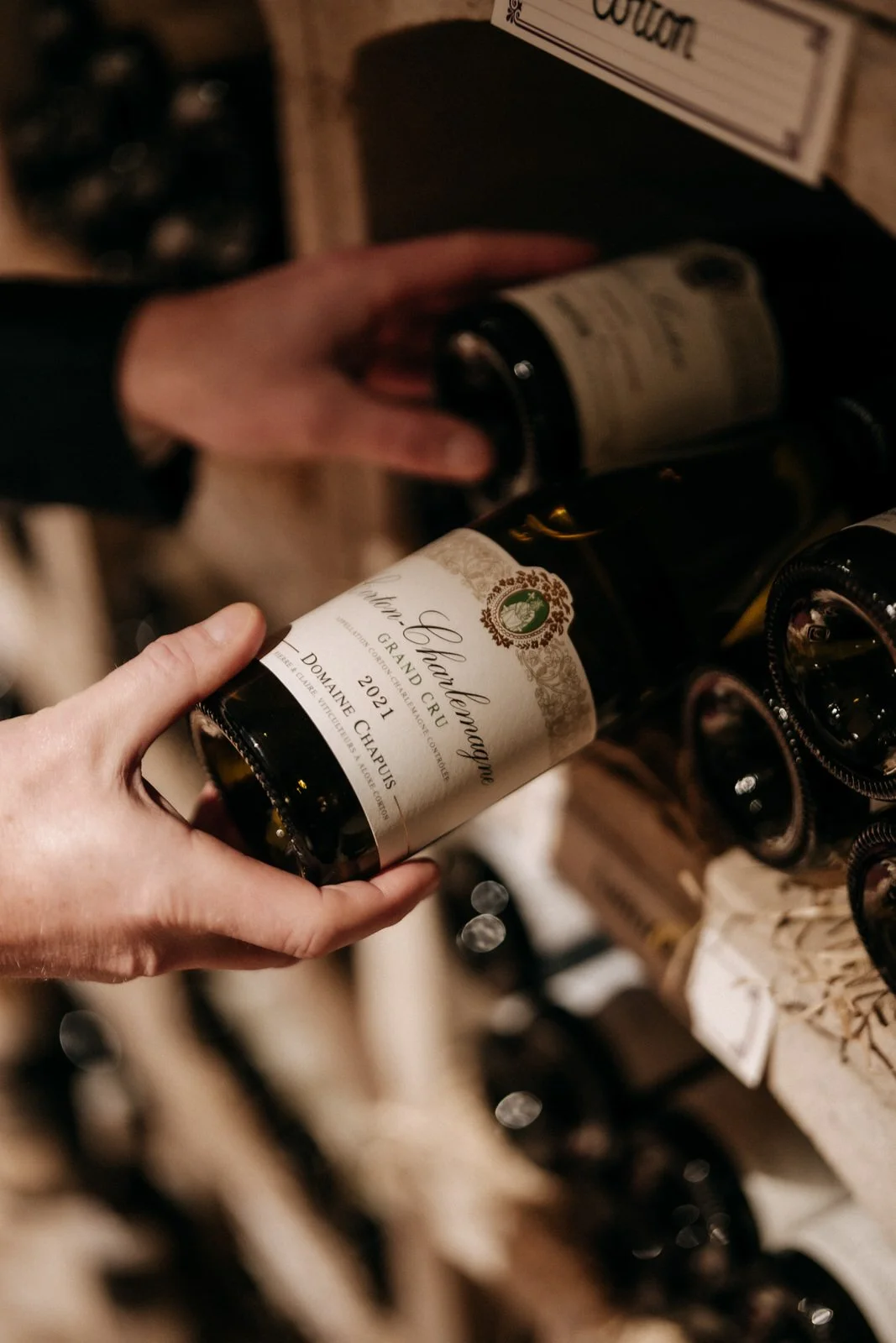 Person holding a bottle of 2021 vintage of Corton-Charlemagne Grand Cru wine in a wine cellar with other bottles on wooden shelves.