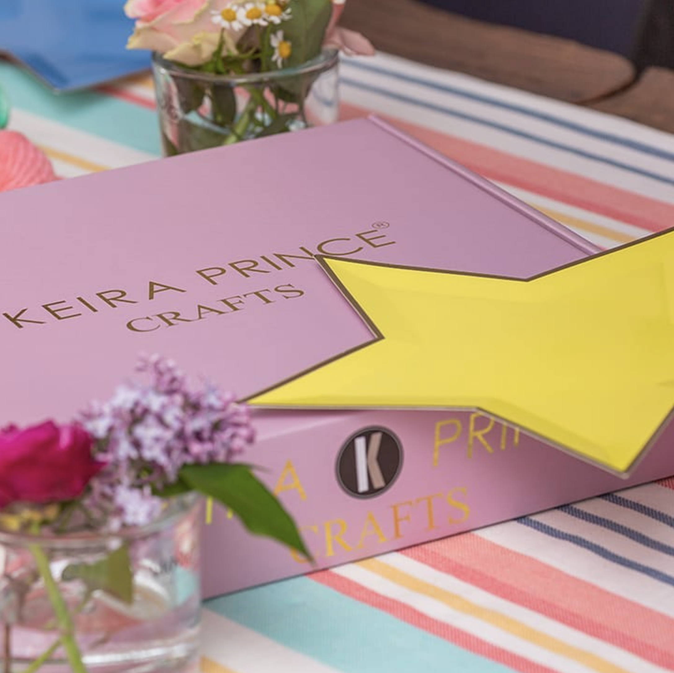 Un mazzo di fiori freschi in un vaso di vetro, posati su una superficie colorata con tessuto a righe in vari colori pastello come rosa, azzurro, giallo e bianco.