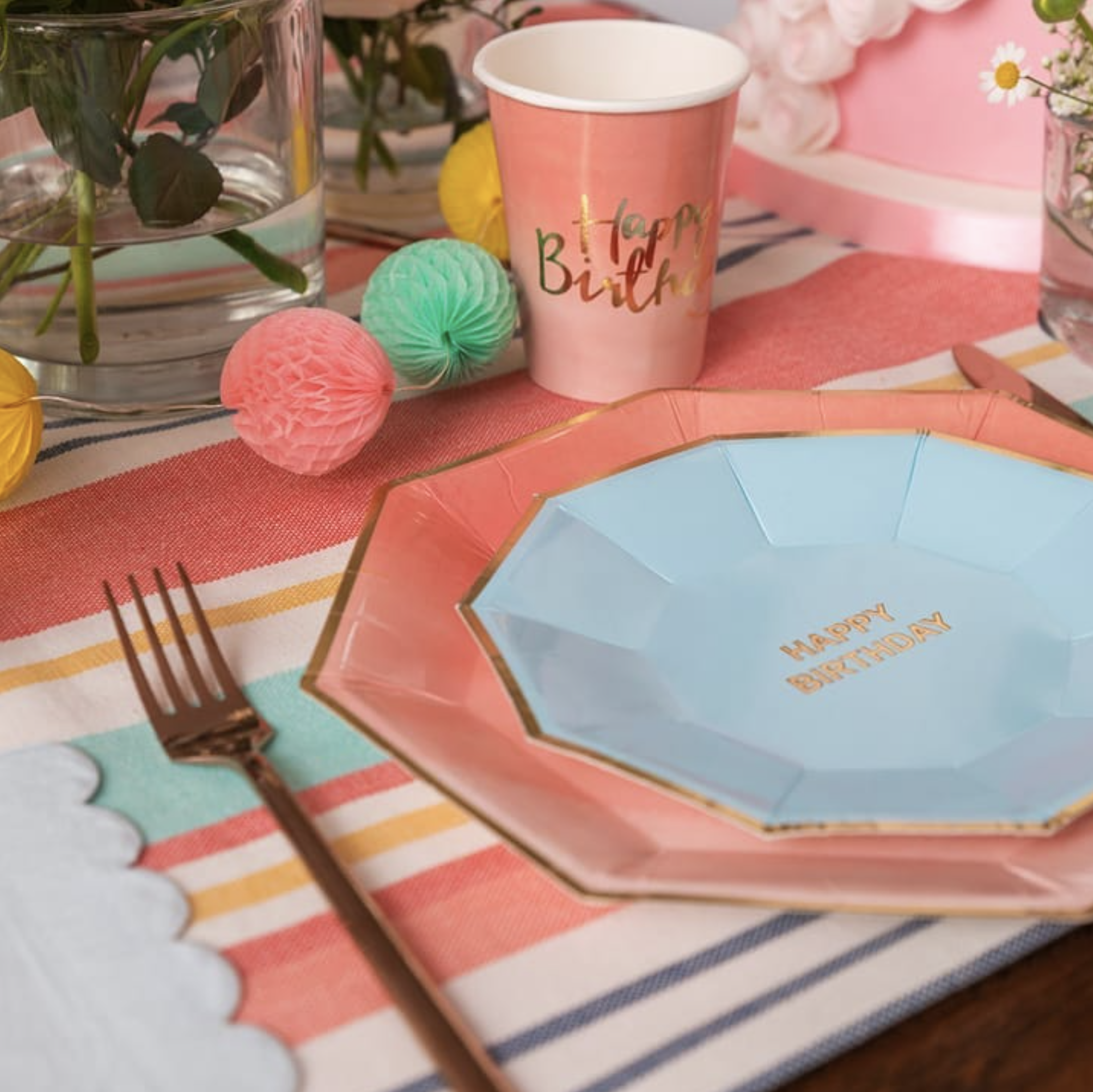 Una festa di compleanno con tovaglioli colorati, bicchieri rosa con scritta "Happy Birthday", decorazioni di carta colorata, e fiori in vasi di vetro.