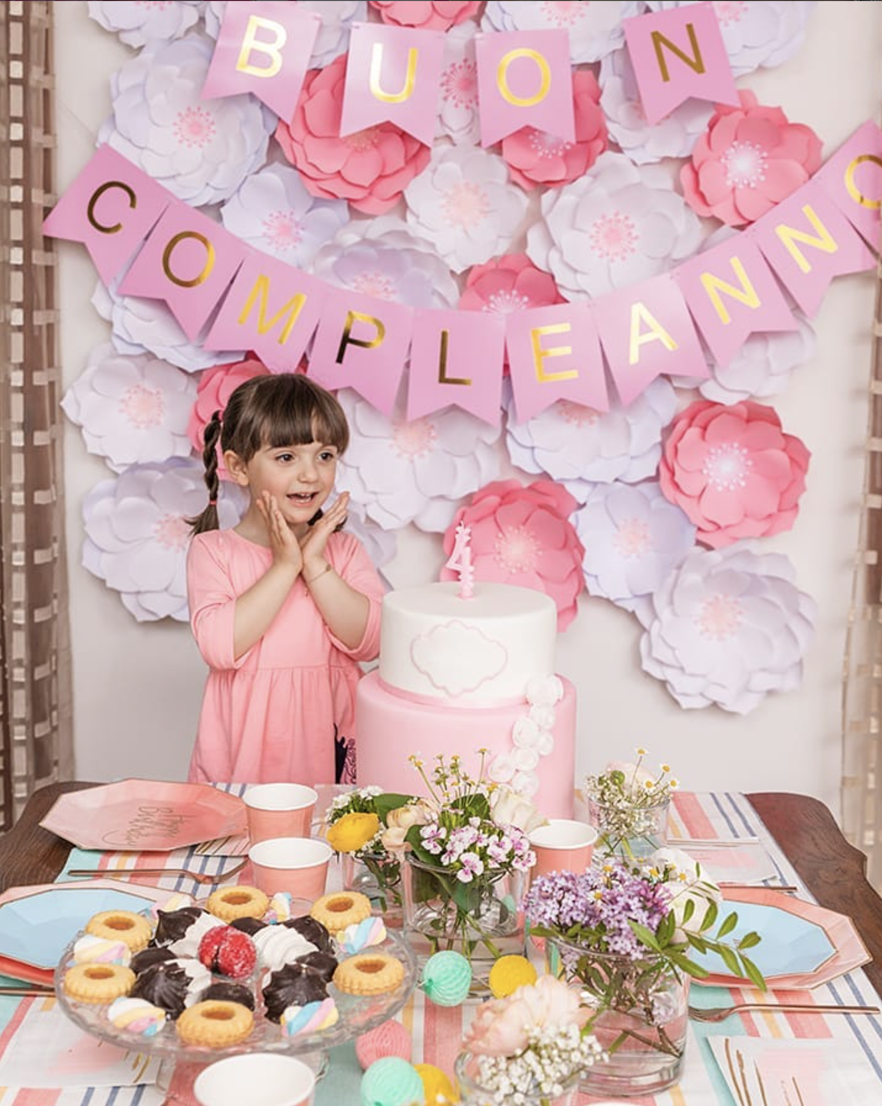 Decorazioni di compleanno con fiori di carta rosa e bianchi, il banner con la scritta 'Buon Compleanno', dolci e fiori sulla tavola.