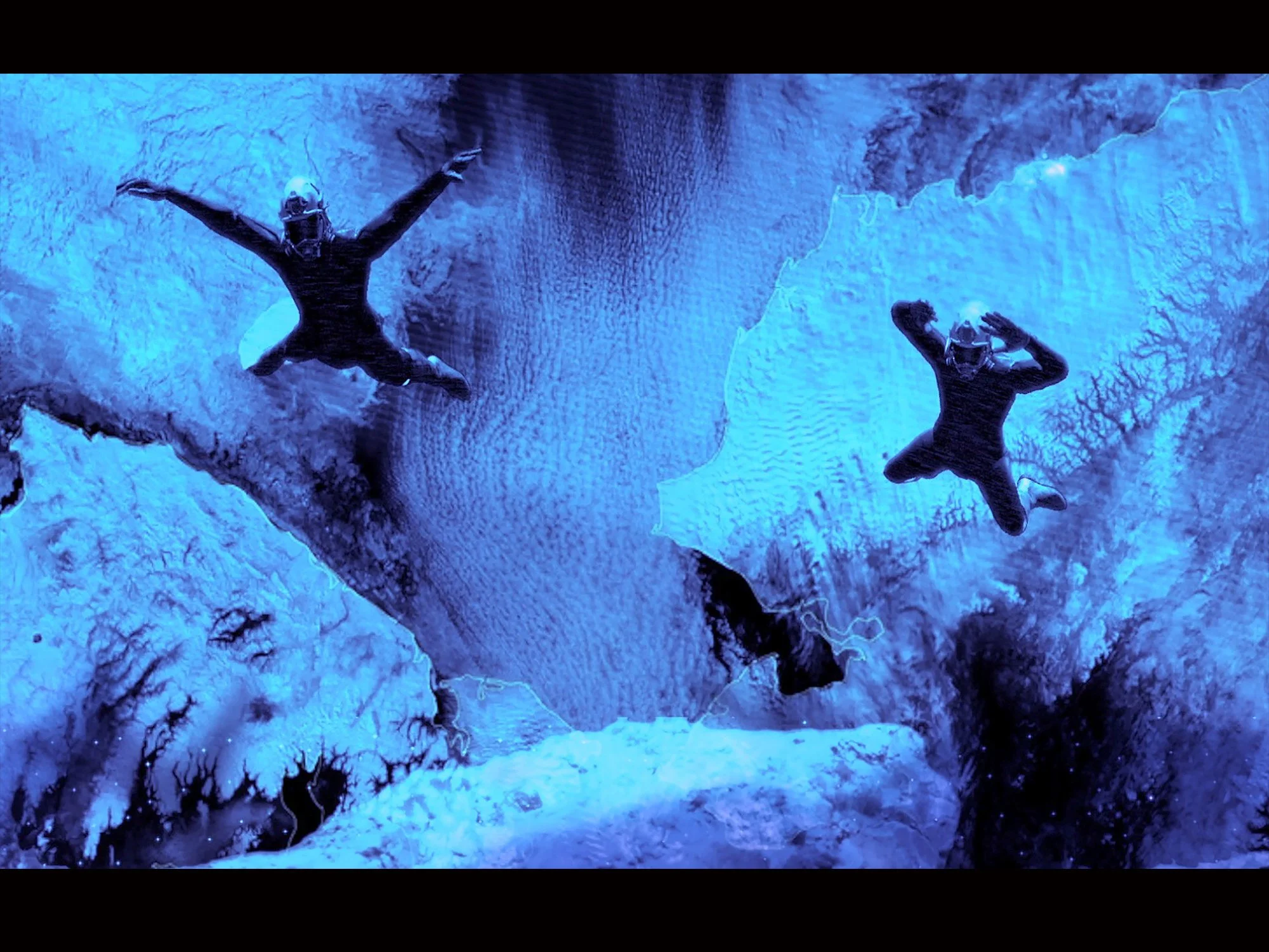 Two divers in black suits and helmets swimming in a blue-lit underwater environment with coral and branching formations.