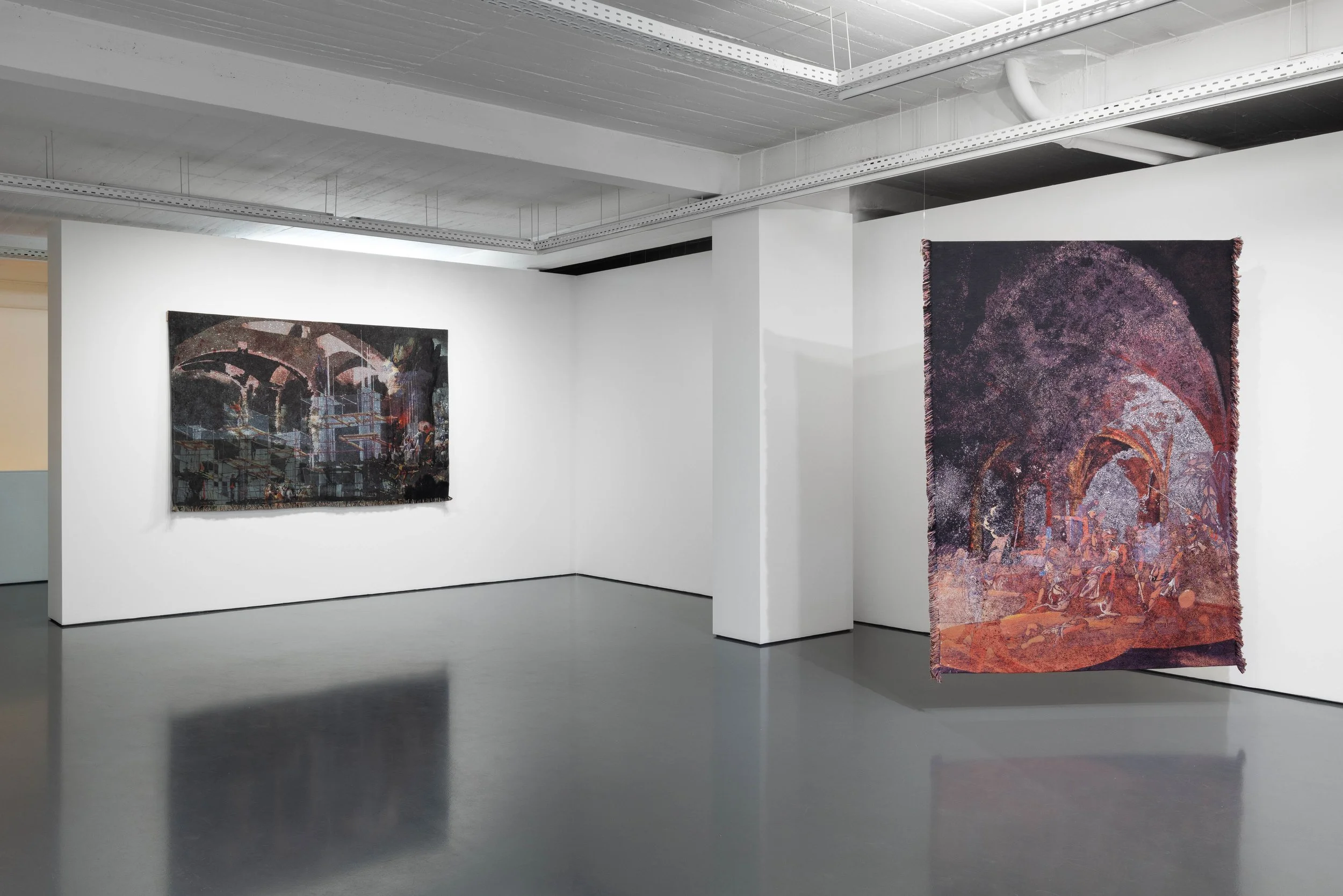 Interior of an art gallery or museum with white walls, displaying two abstract tapestries hanging on the walls, one on the left with dark colors and the other on the right with shades of purple, pink, and purple. The floor is gray, and the ceiling is industrial with visible pipes and wiring.