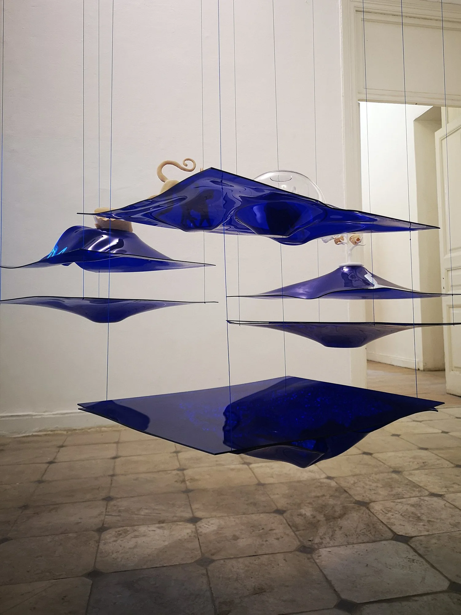 An art installation of hanging glass sculptures in shades of blue and clear, arranged in a layered formation, hanging in a gallery with tiled floors and a white wall.