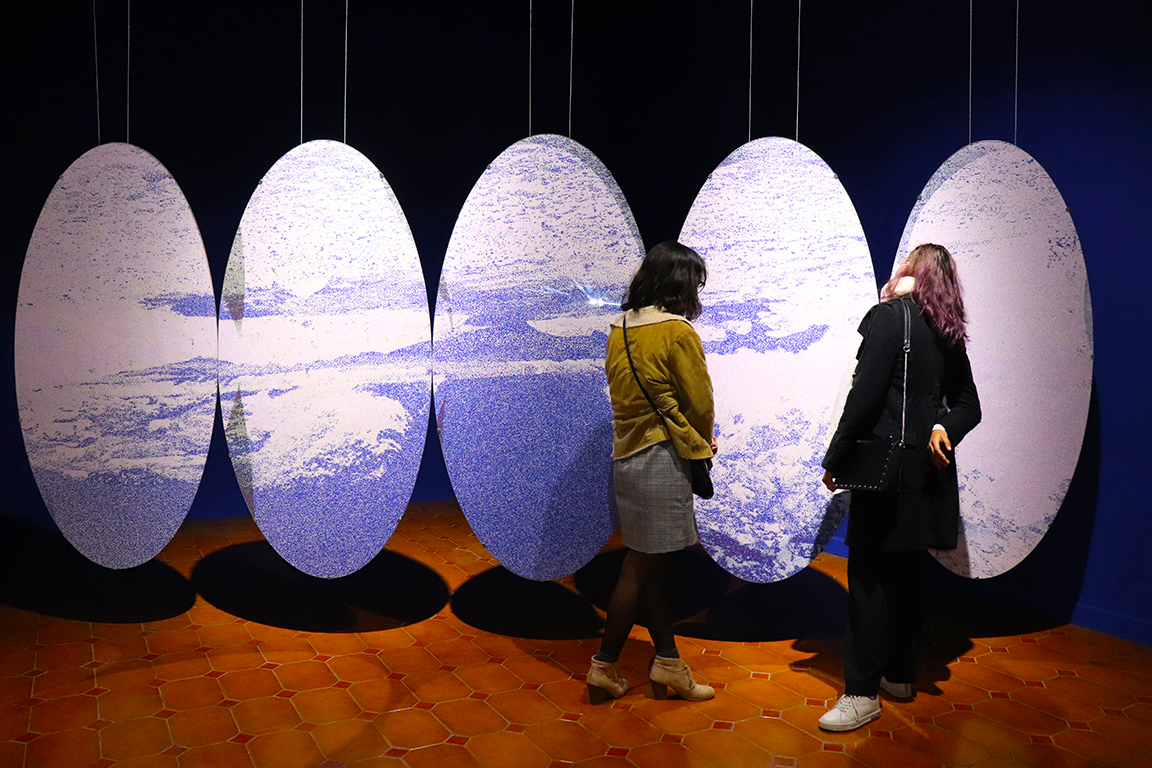 Two women standing and looking at an art installation featuring six oval mirrors reflecting clouds and sky, with a brick floor and dark background.