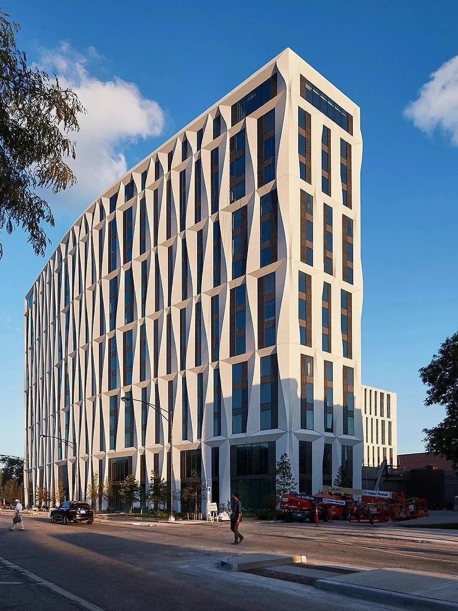Modern high-rise building with a unique geometric facade, located on an urban street with trees, vehicles, and pedestrians, under a blue sky with clouds.