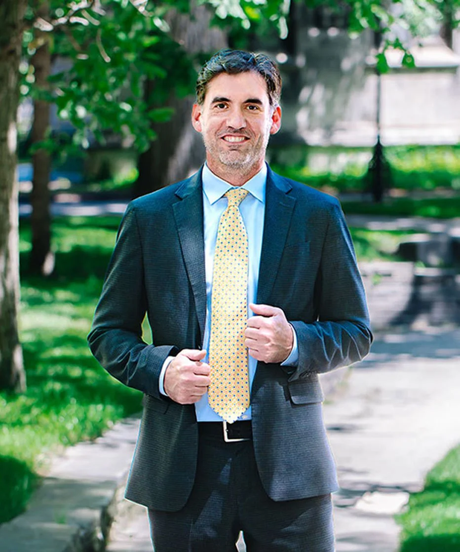 A man in a suit and yellow tie standing outdoors on a sidewalk surrounded by trees and greenery, smiling at the camera.