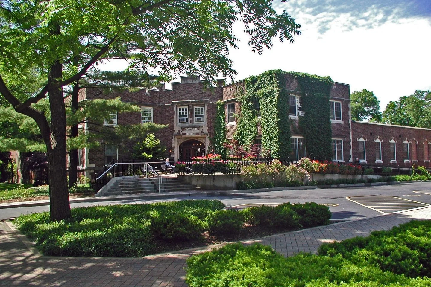 Historic brick building labeled Scottish Lodge, surrounded by greenery, ivy climbing the walls, a small garden with flowers in front, and a parking lot nearby.