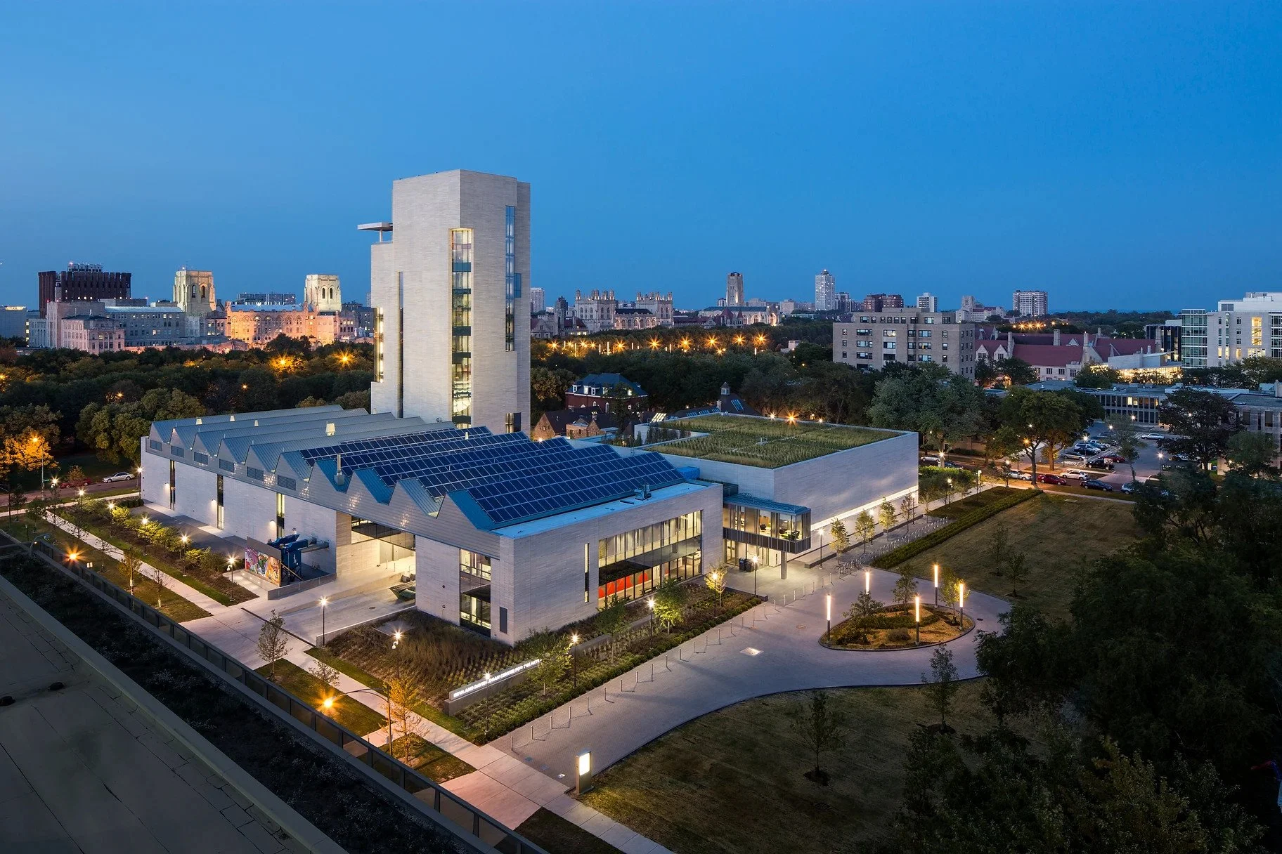 A modern cityscape at twilight featuring a contemporary building with solar panels on its roof, surrounded by trees, walkways, and city lights with a mix of historic and modern architecture in the background.