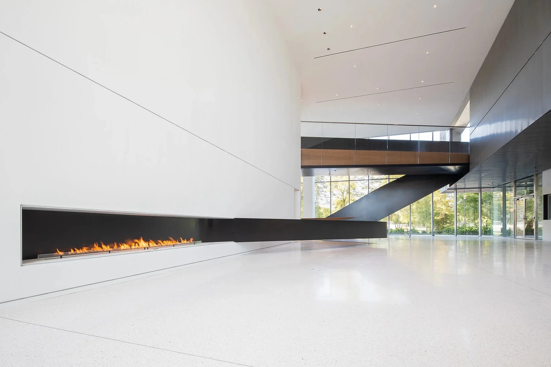 Modern interior with a long, horizontal fireplace on a white wall, large floor-to-ceiling windows, an upstairs balcony with a glass railing, and a staircase leading to the second floor. Outside visible greenery.