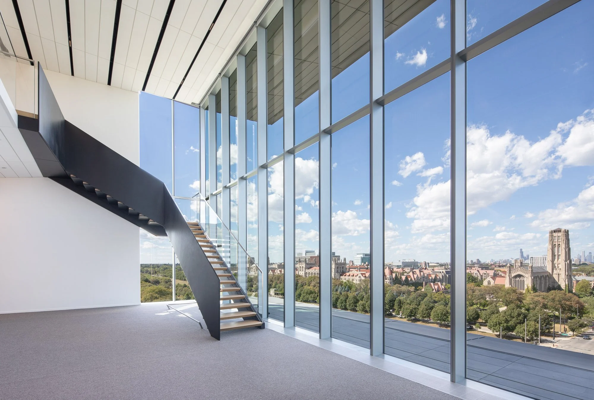 Interior of a modern building with large glass windows and a staircase leading to a higher level, overlooking a cityscape with church towers, green trees, and blue sky with clouds.