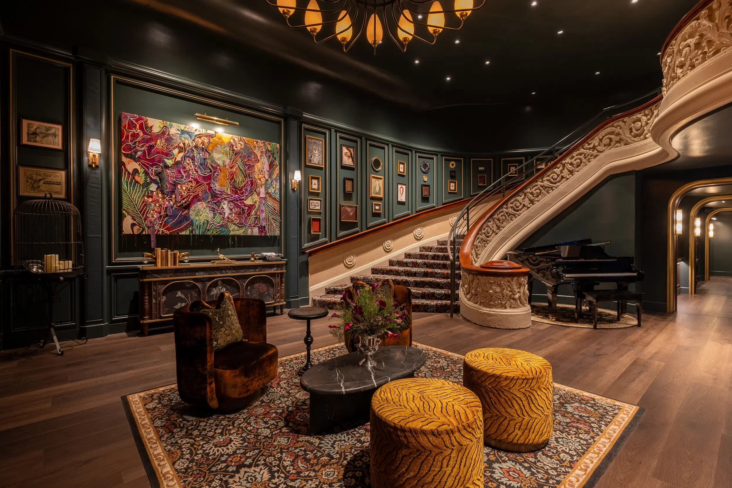 A luxurious interior with a grand staircase, ornate detailing, and dark walls decorated with framed artwork. There's a large colorful tapestry on the wall and inviting seating including velvet armchairs and a patterned rug. A chandelier hangs from the ceiling and a grand piano is partially visible beneath the staircase.