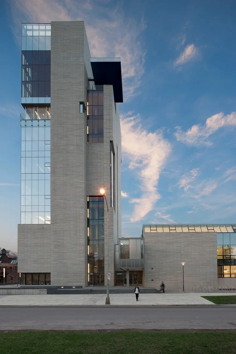 Modern multi-story building with large glass windows and concrete exterior, under a partly cloudy sky, with a person walking on the sidewalk in front.