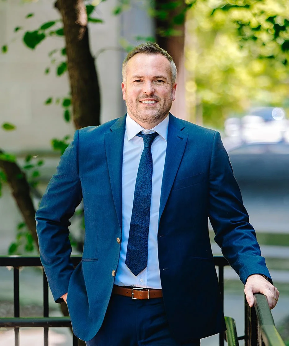 A man in a blue suit, white shirt, and blue tie standing outdoors on a balcony with greenery and trees in the background, smiling at the camera.