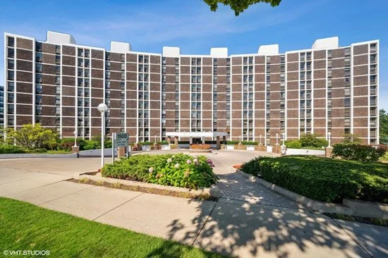 A large curved brick apartment building with white accents, surrounded by landscaped lawns and walkways with flowers and bushes, under a blue sky.