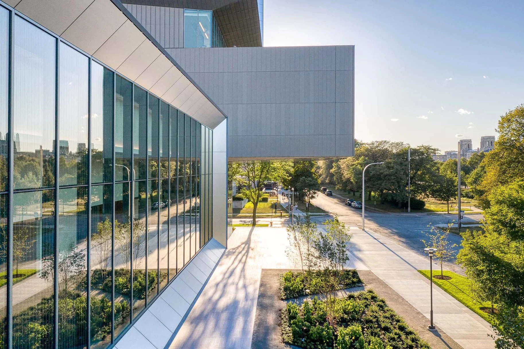 Modern glass building with reflective windows, surrounding trees, a wide sidewalk, and a street with parked and moving cars, with city skyline in the background under a clear sky.