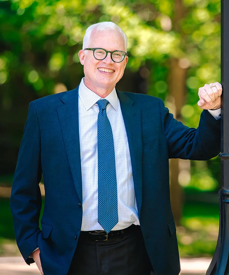 Smiling man in a navy suit, light blue shirt, and dotted tie, wearing glasses, outdoors with green trees in the background, standing next to a post.