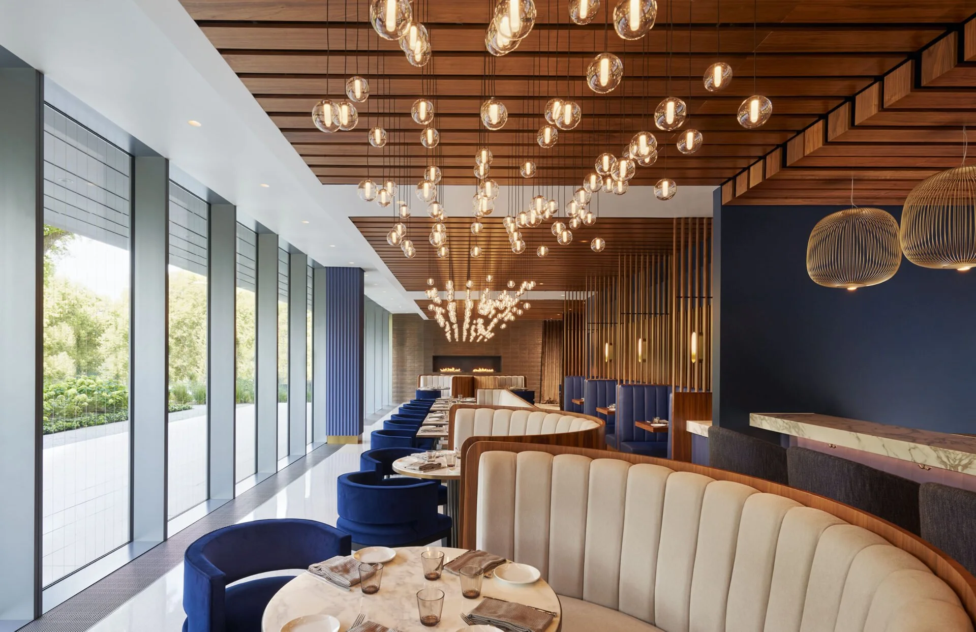 Interior of a modern restaurant with large windows, wooden ceiling with hanging glass light fixtures, blue and beige upholstered seats around marble tables, and a dark blue accent wall.