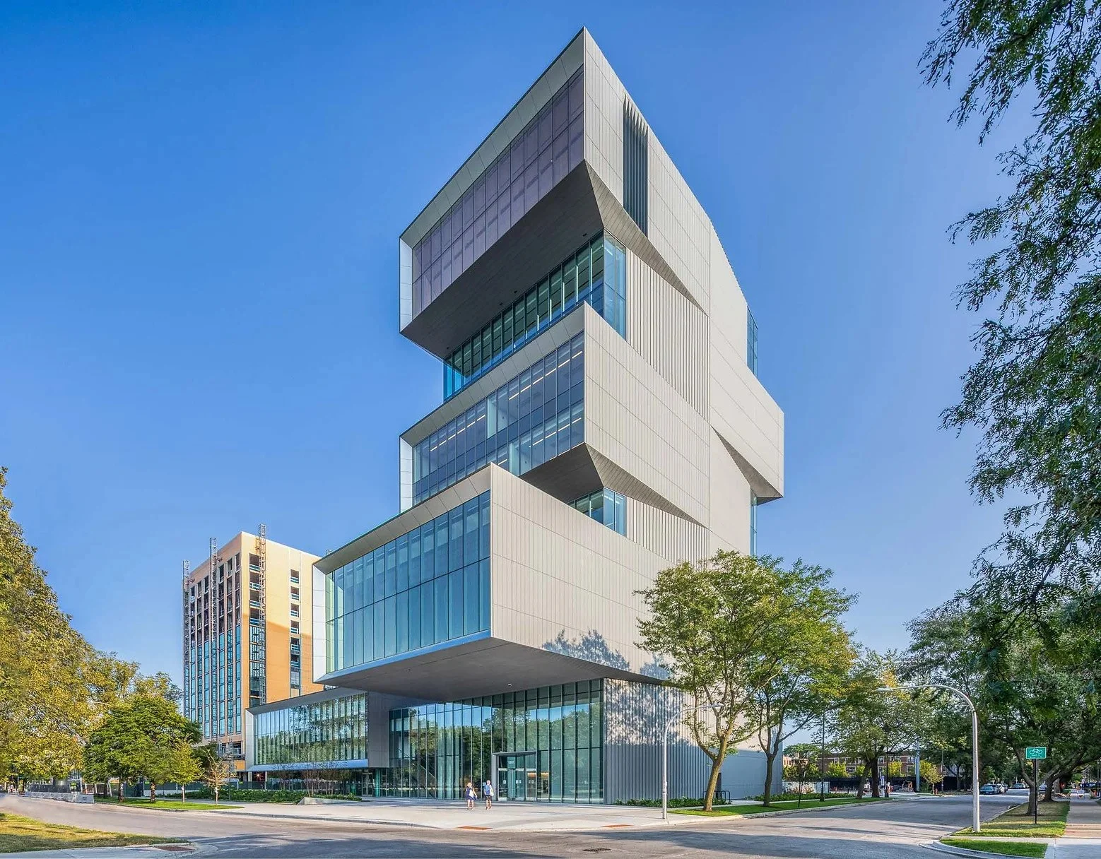 Modern multi-story building with stacked glass and metal sections, surrounded by trees and a sidewalk.