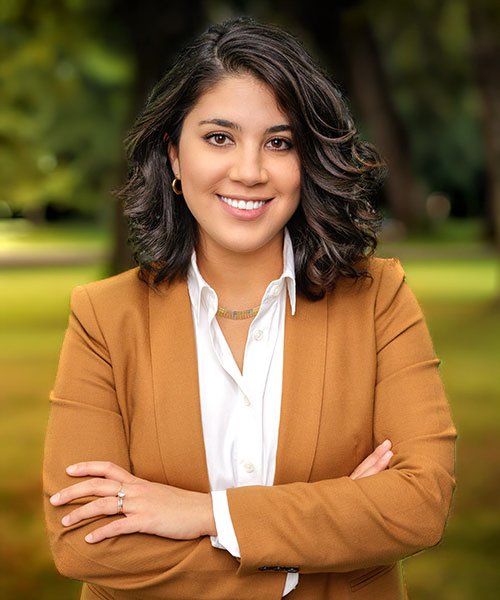 A young woman with shoulder-length dark hair, wearing a tan blazer over a white blouse, smiling while standing outdoors in a park.
