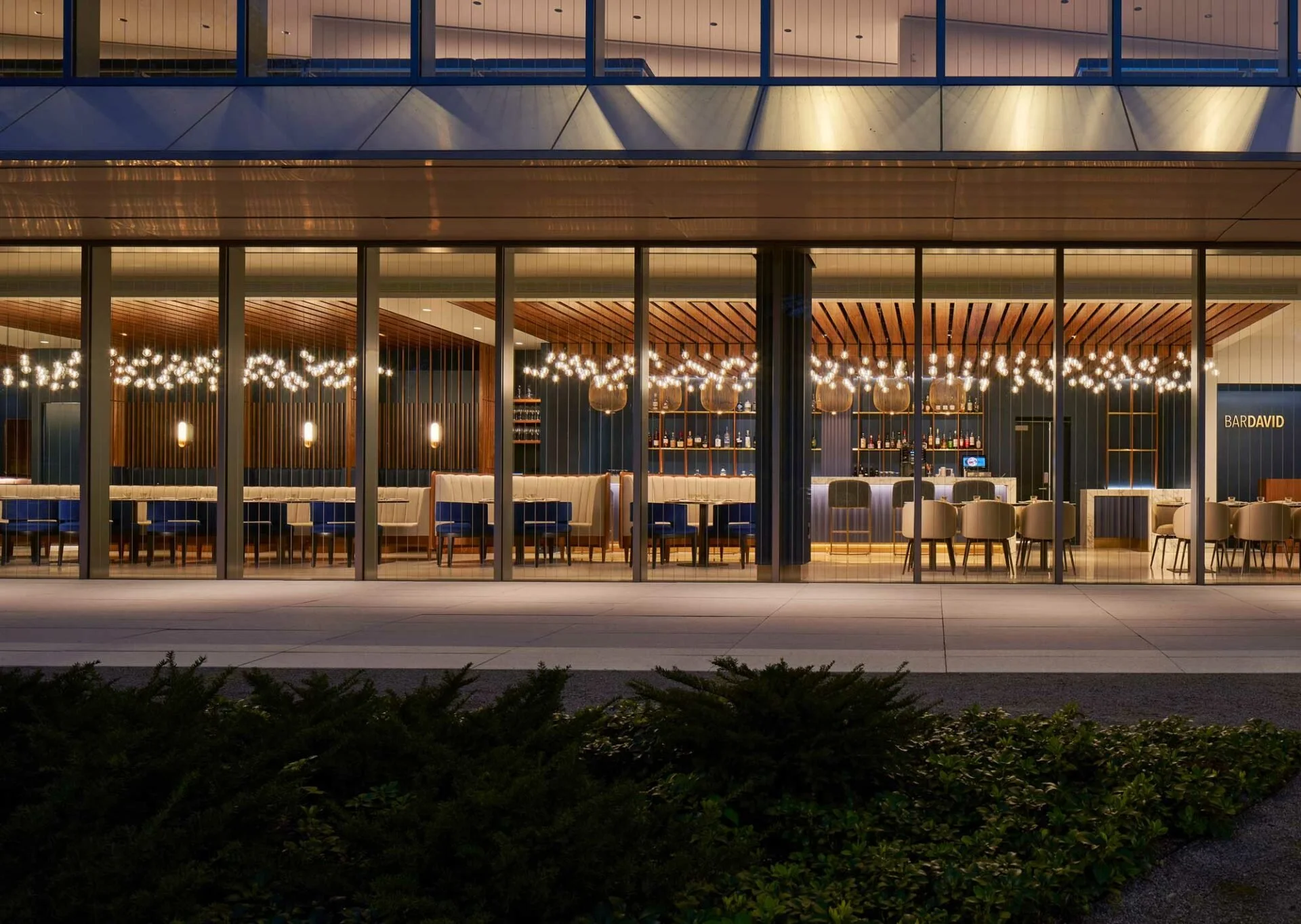 Night view of a modern restaurant with glass exterior, interior lighting, wooden accents, and a bar with chairs.