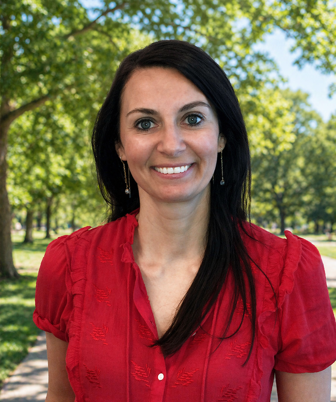 A woman with long dark hair, blue eyes, and earrings, smiling outdoors in a park with green trees in the background, wearing a red blouse.