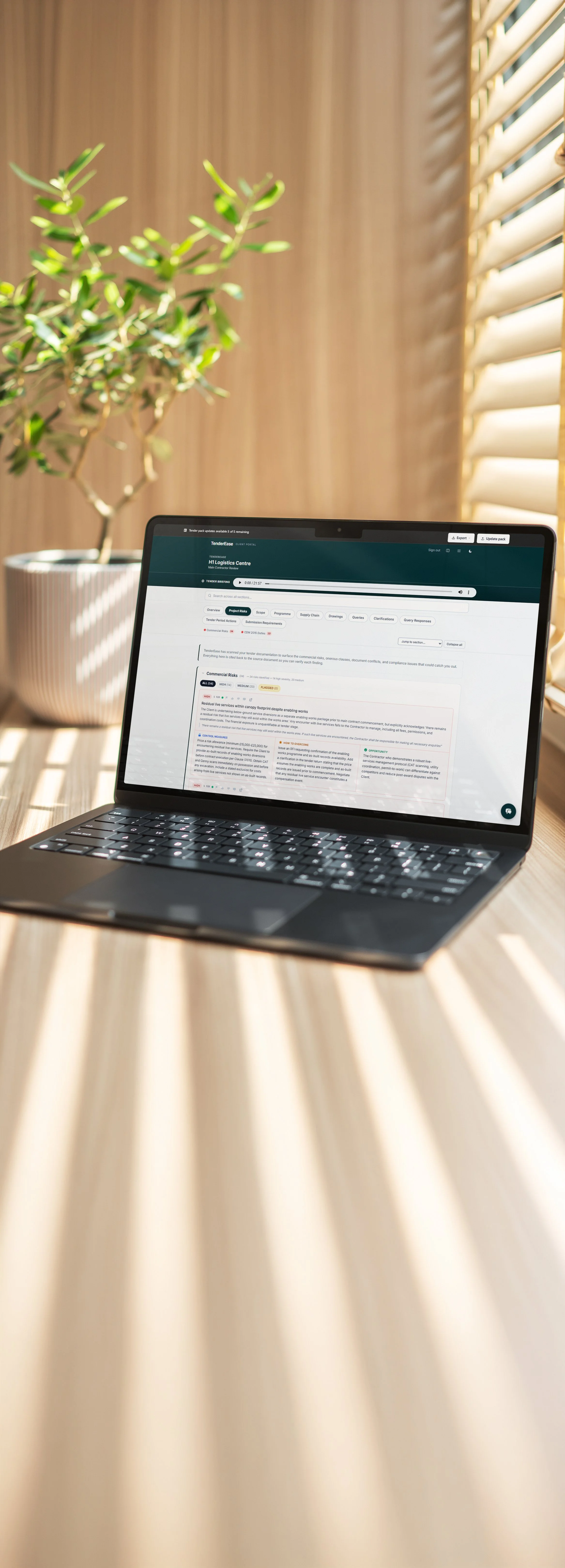 Laptop on a wooden desk displaying a webpage with a green header, beside a potted plant with green leaves, sunlight coming through window blinds.