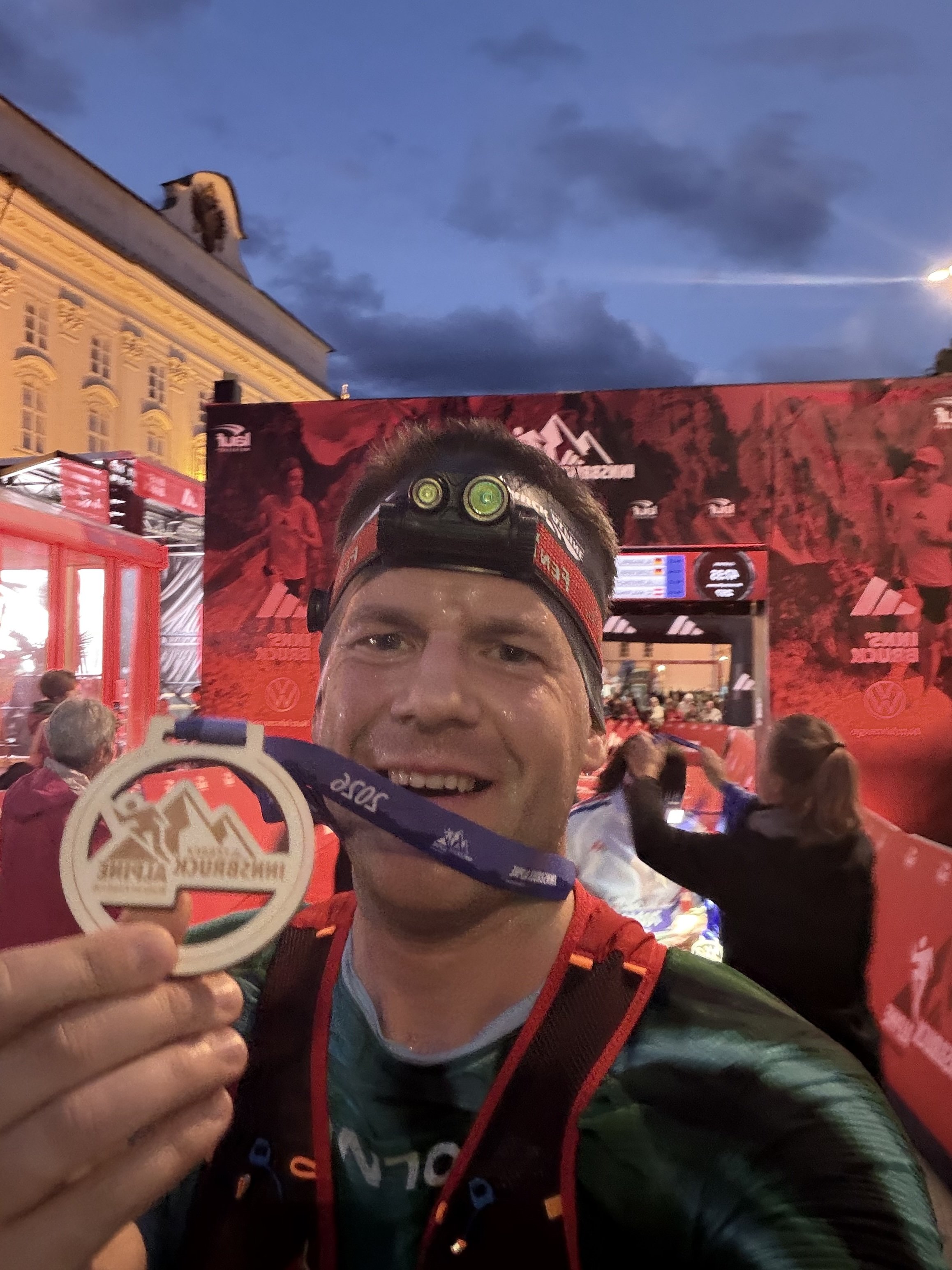 Ein Mann in Laufkleidung hält eine Finisher-Medaille und lächelt bei einer Laufveranstaltung am Abend, im Hintergrund sind andere Teilnehmer und eine große Bühne zu sehen.