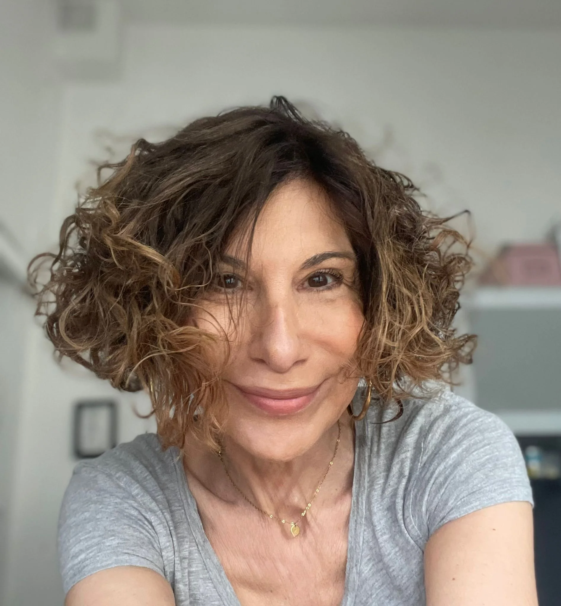 Une femme souriante avec des cheveux bouclés et bruns, portant un t-shirt gris clair et un collier doré, prenant un selfie dans un intérieur.