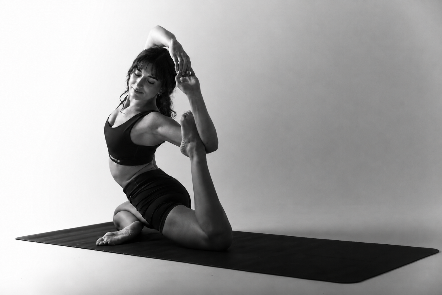 Une femme pratique le yoga sur un tapis, exécutant une posture d'étirement en position genouillère, en noir et blanc.