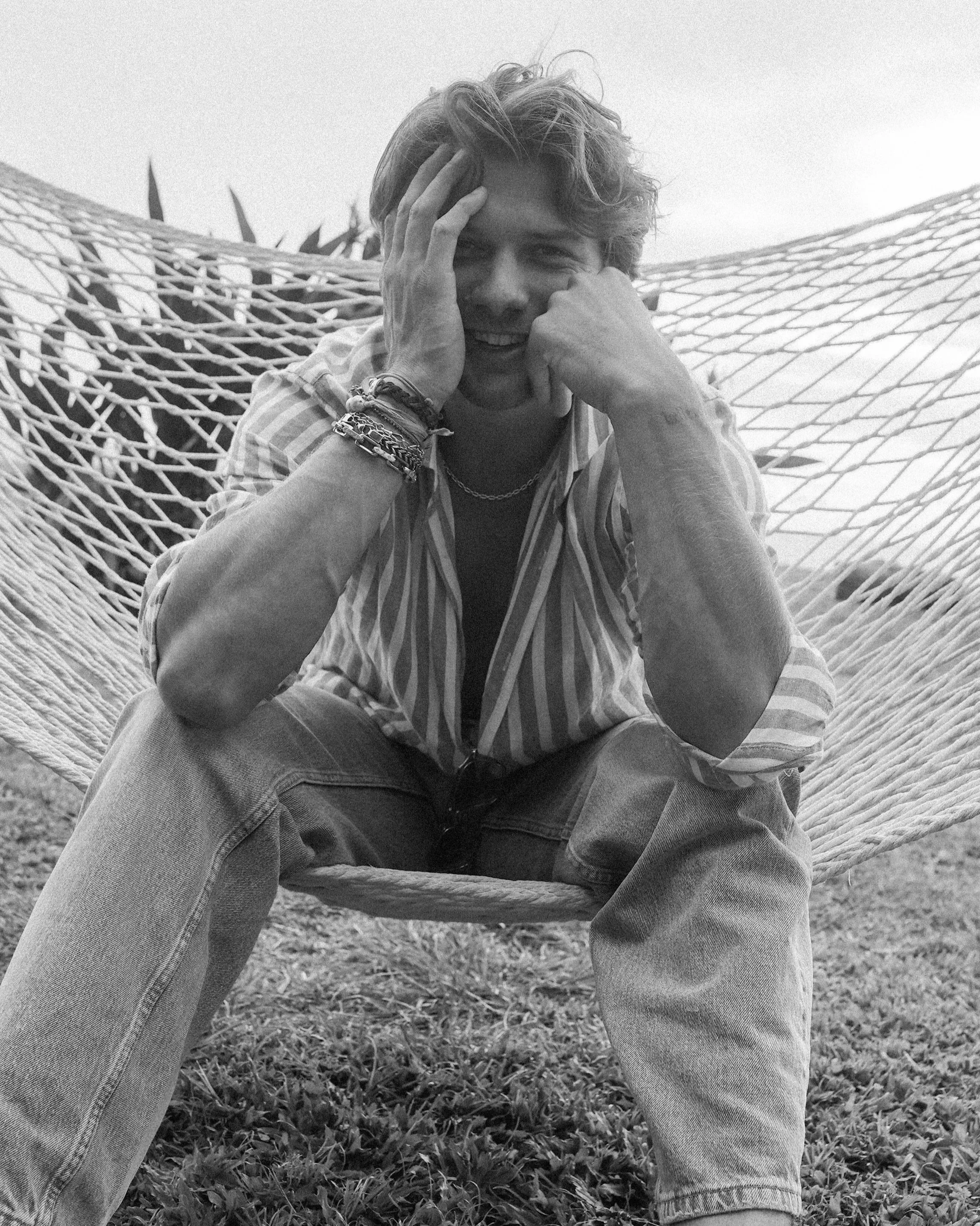 Une personne assise dans une balançoire en corde en plein air, portant un vêtement à rayures. La personne a les cheveux courts et bouclés, et porte plusieurs bracelets et un collier. La photo est en noir et blanc.