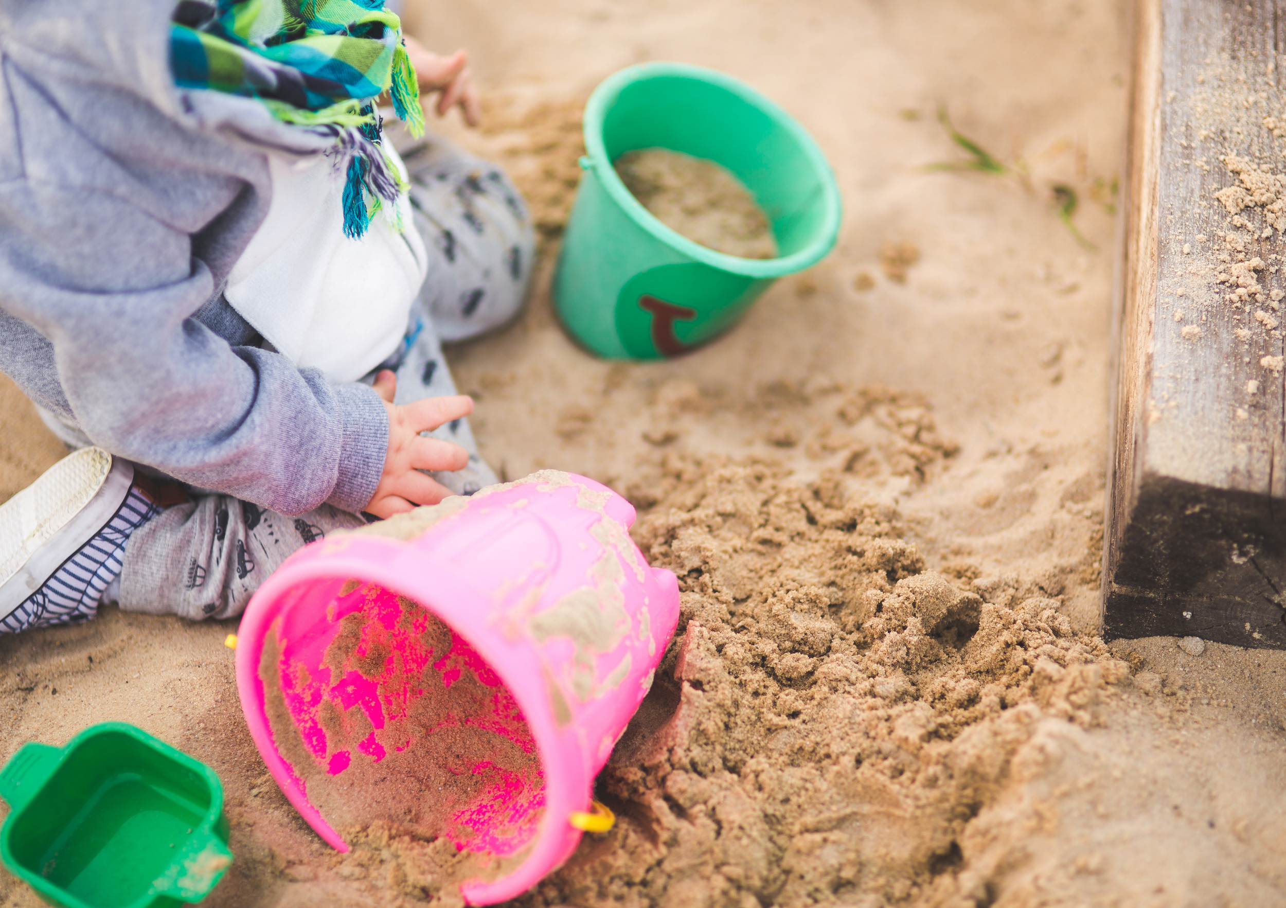 Child playing in sand, with colorful plastic buckets and shovels, wearing a gray hoodie and a multicolored scarf.