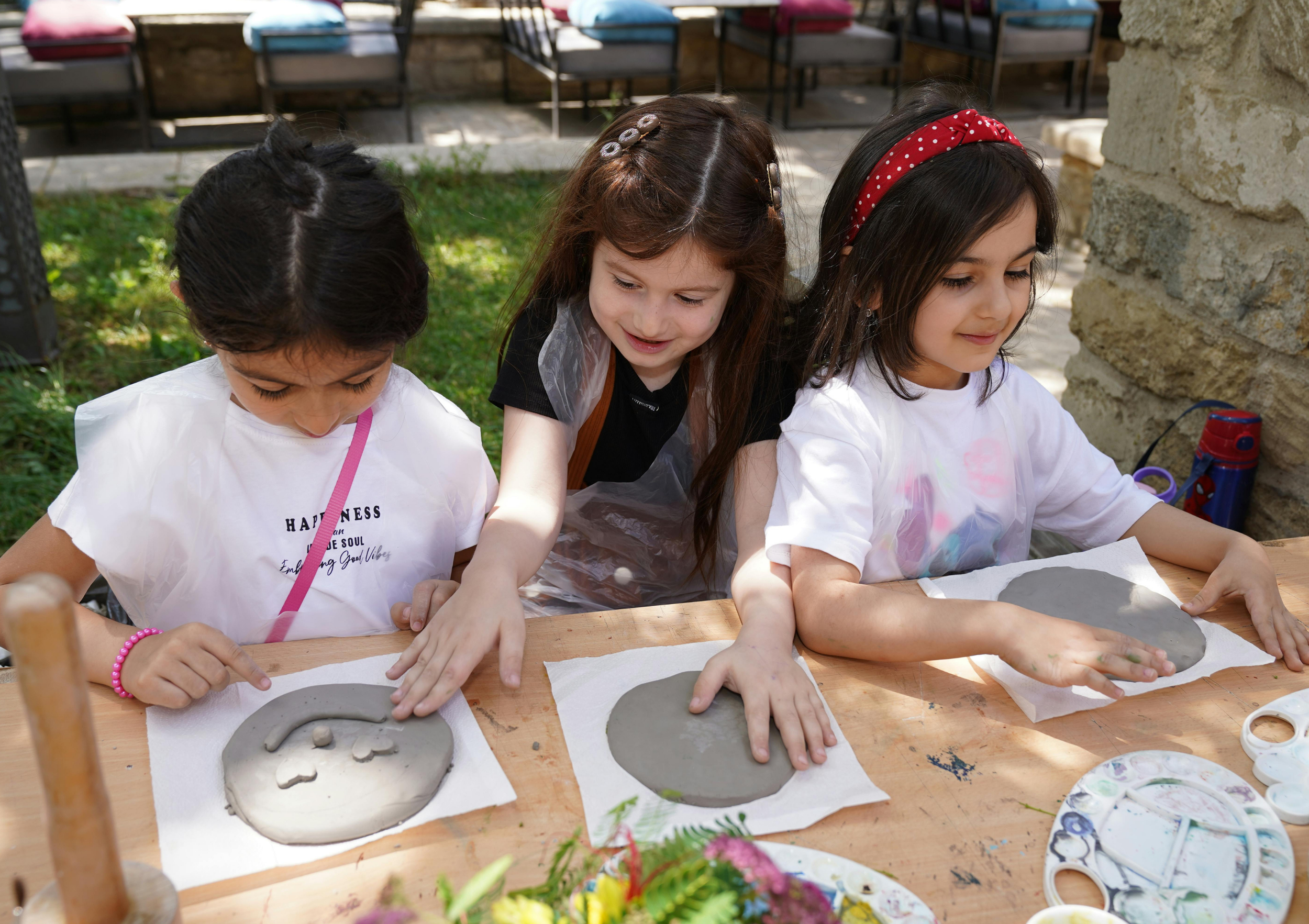Three young girls are sitting at a wooden table outdoors, working on clay art projects. They are focused on shaping the clay in front of them, with art supplies and a colorful bouquet of flowers visible on the table.