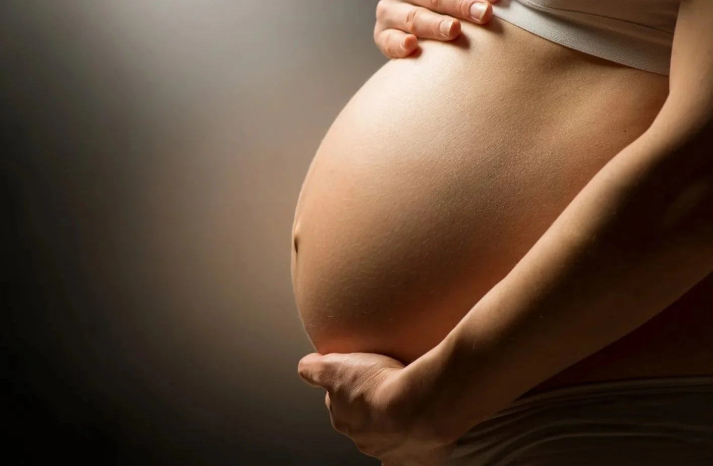 Une femme enceinte tient son ventre arrondi avec une main. La photo met en valeur son ventre et ses mains, avec un fond neutre.