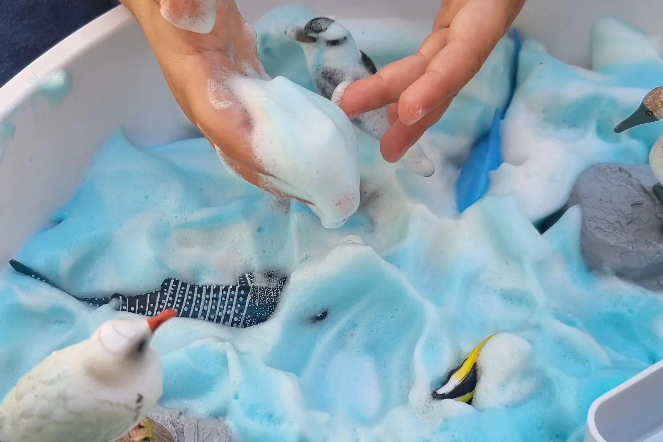 des mains d'enfants entrain de jouer avec de l'aquafaba et du colorant alimentaire bleu pour faire l'océan avec des animaux marin