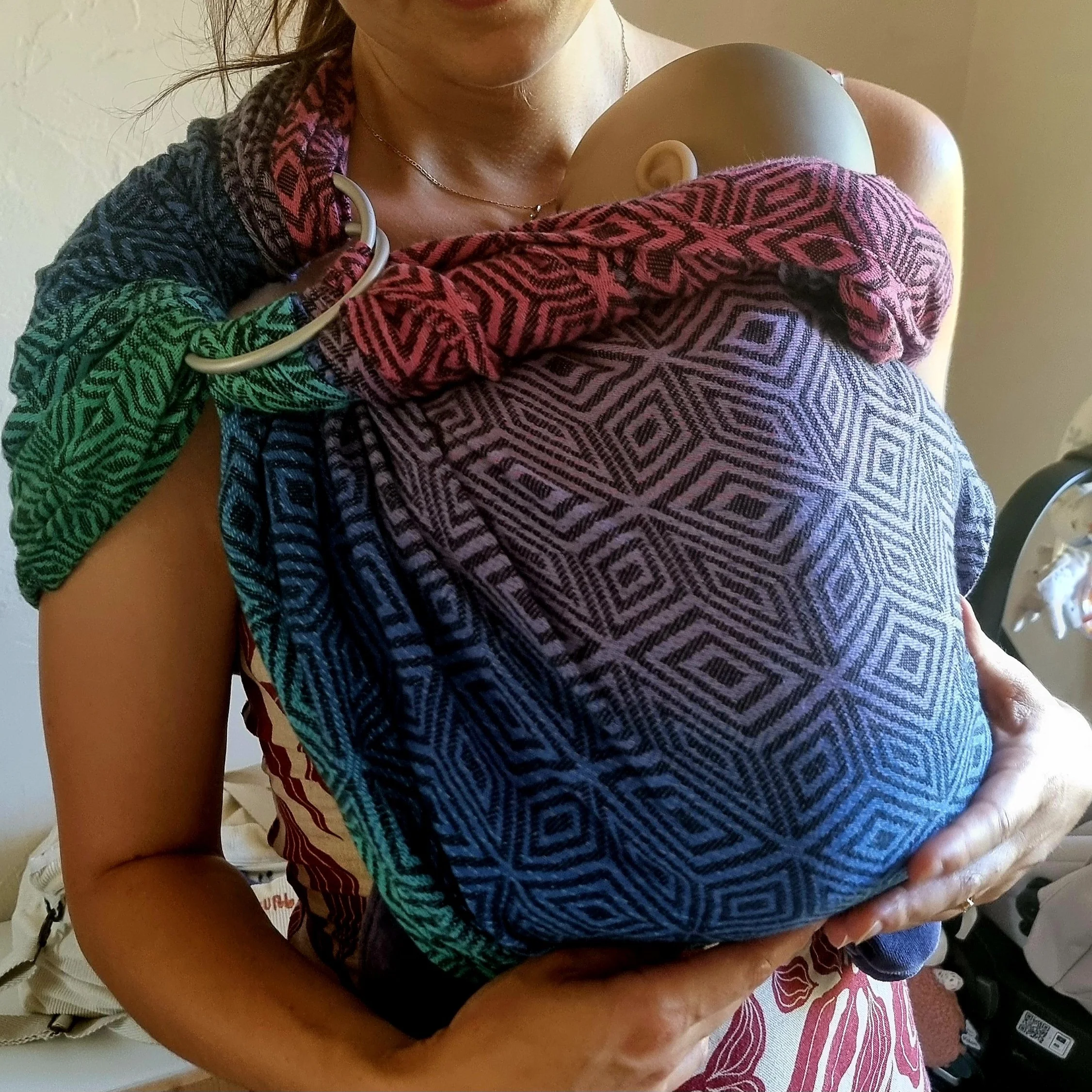 Femme portant un bébé avec un sling lors d'un atelier de portage avec une monitrice de portage. Le sling est multicolore et la femme soutien son bébé par le siège.