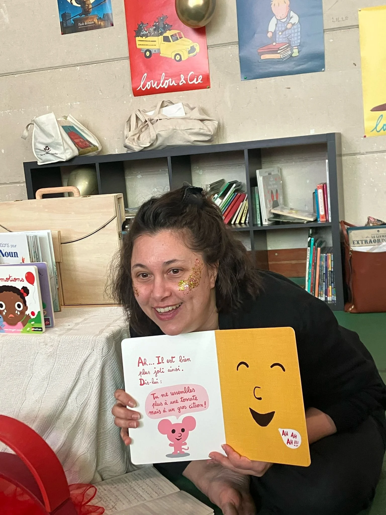 Une femme tient un livre pour enfants avec un visage souriant et un petit ours rose illustré à l'intérieur. Elle sourit et a des paillettes dorées sur la joue. La photo est prise dans une salle de classe, avec des étagères remplies de livres en arrière-plan et quelques posters colorés au mur.