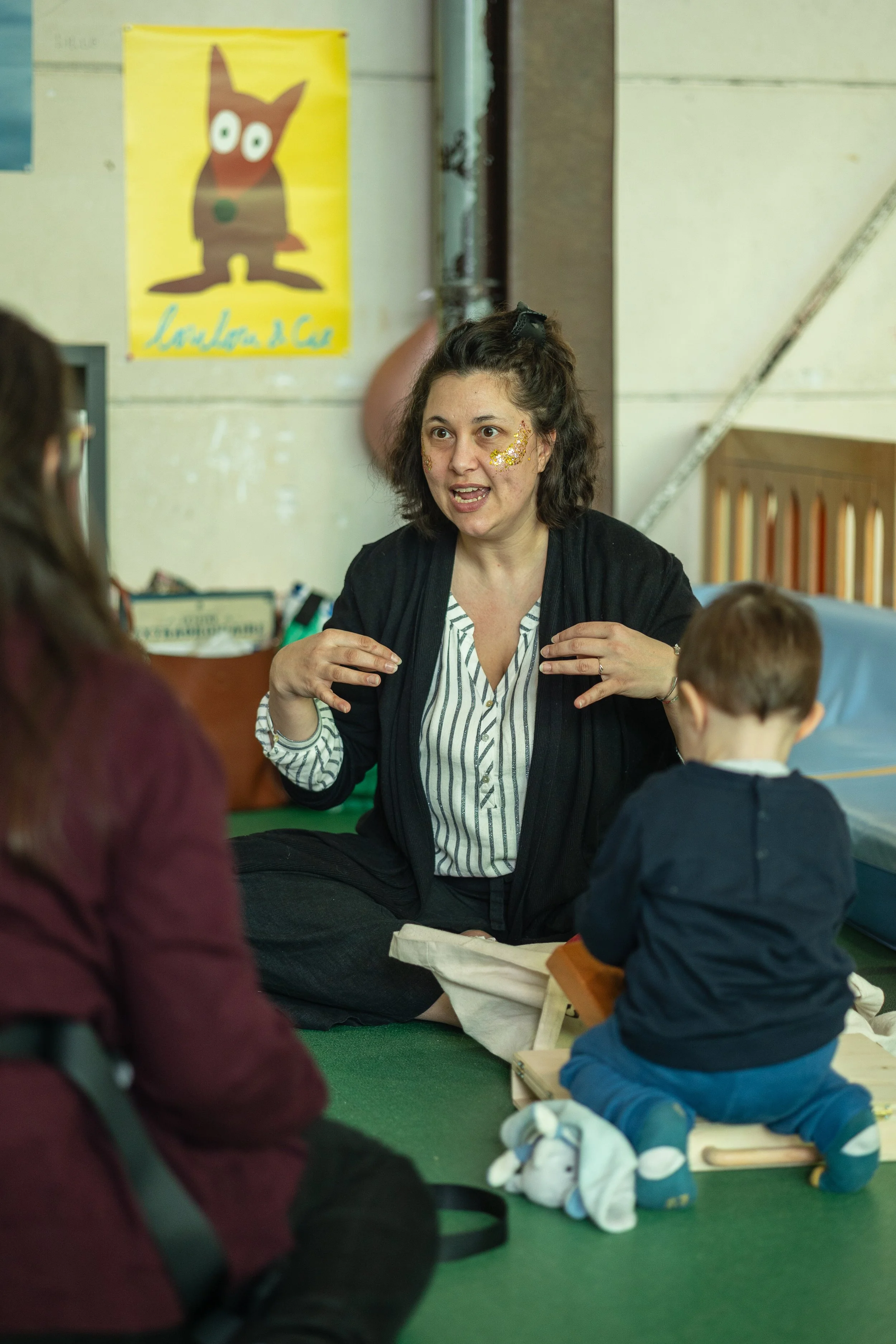 Une femme parle à un groupe d'enfants assis sur le sol, dans une salle de classe. La femme a des cheveux bruns bouclés et a des paillettes dorées sur le visage. Elle est entrain de signer une histoire en LSF