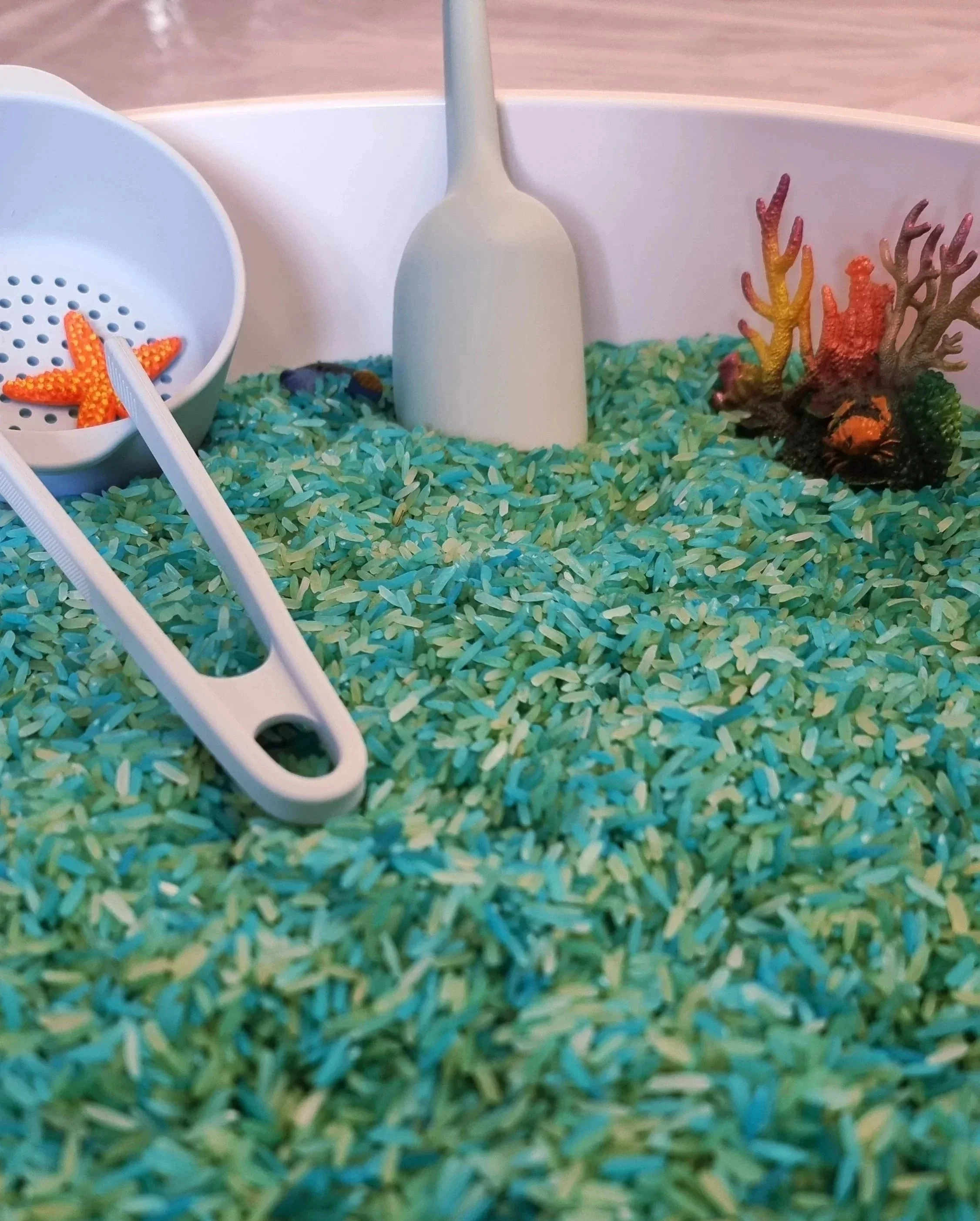 On voit un mélange de riz bleu et vert claire avec du matériel de transvasement et de petits animaux de la mer pour un bac sensoriel "cherche et trouve"