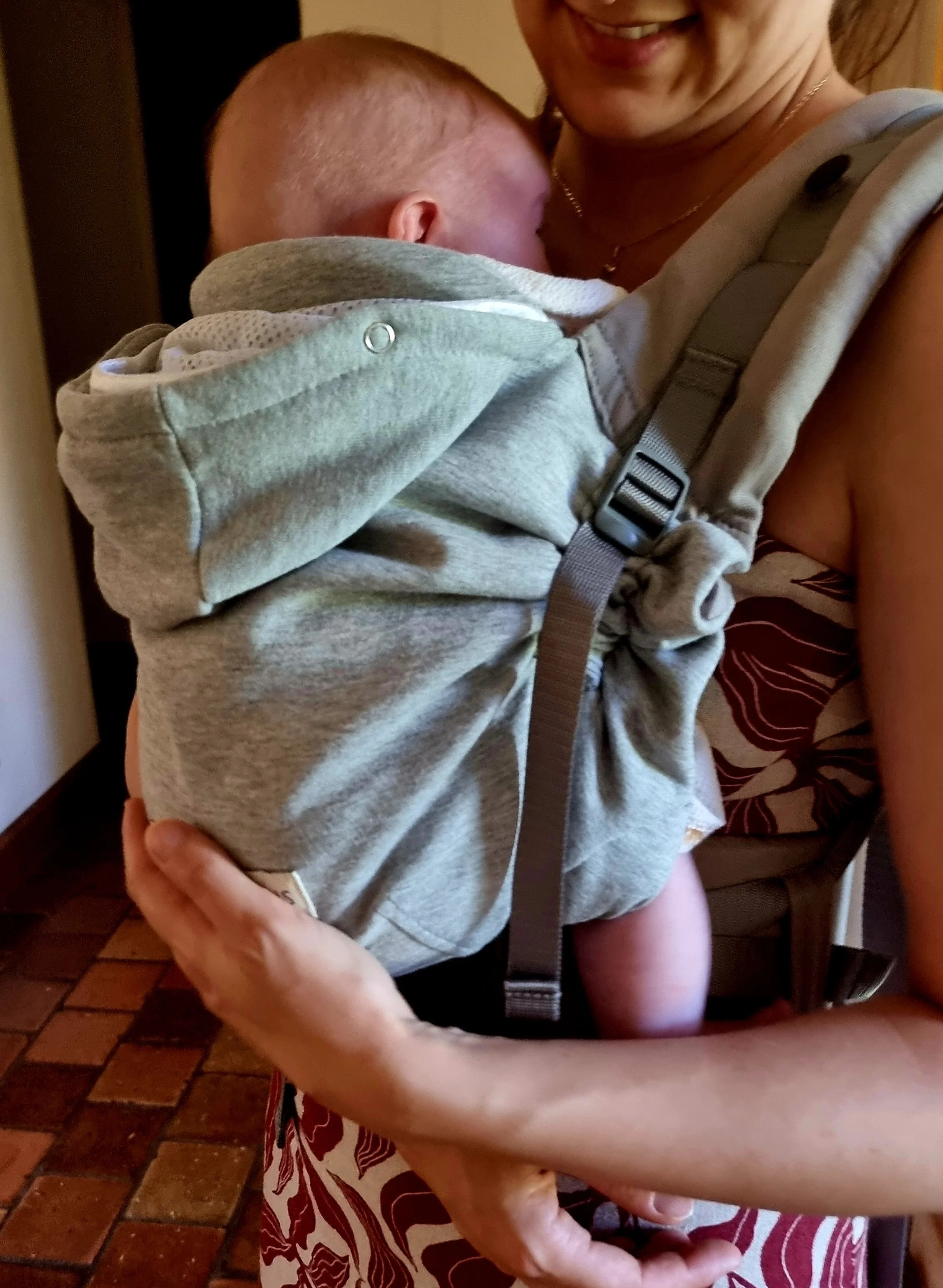 Une femme porte son bébé dans un porte bébé préformé lors d'un atelier de portage avec une monitrice. Le porte bébé est gris et la femme passe un moment doux avec son bébé.