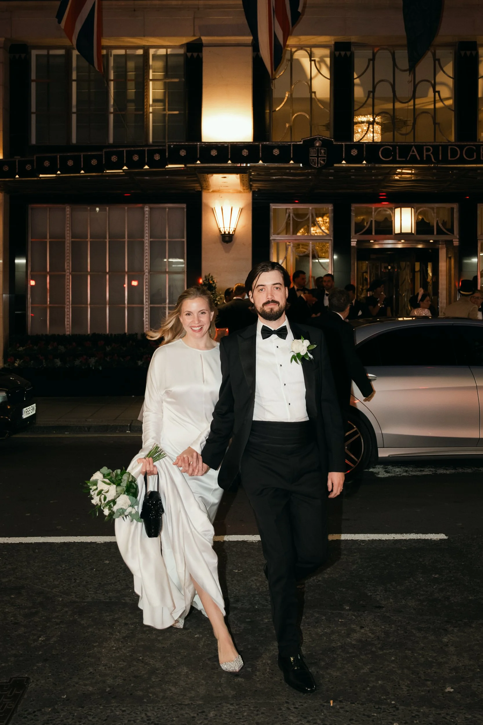 Bride and groom walking past Claridge's at night, the hotel lit behind them